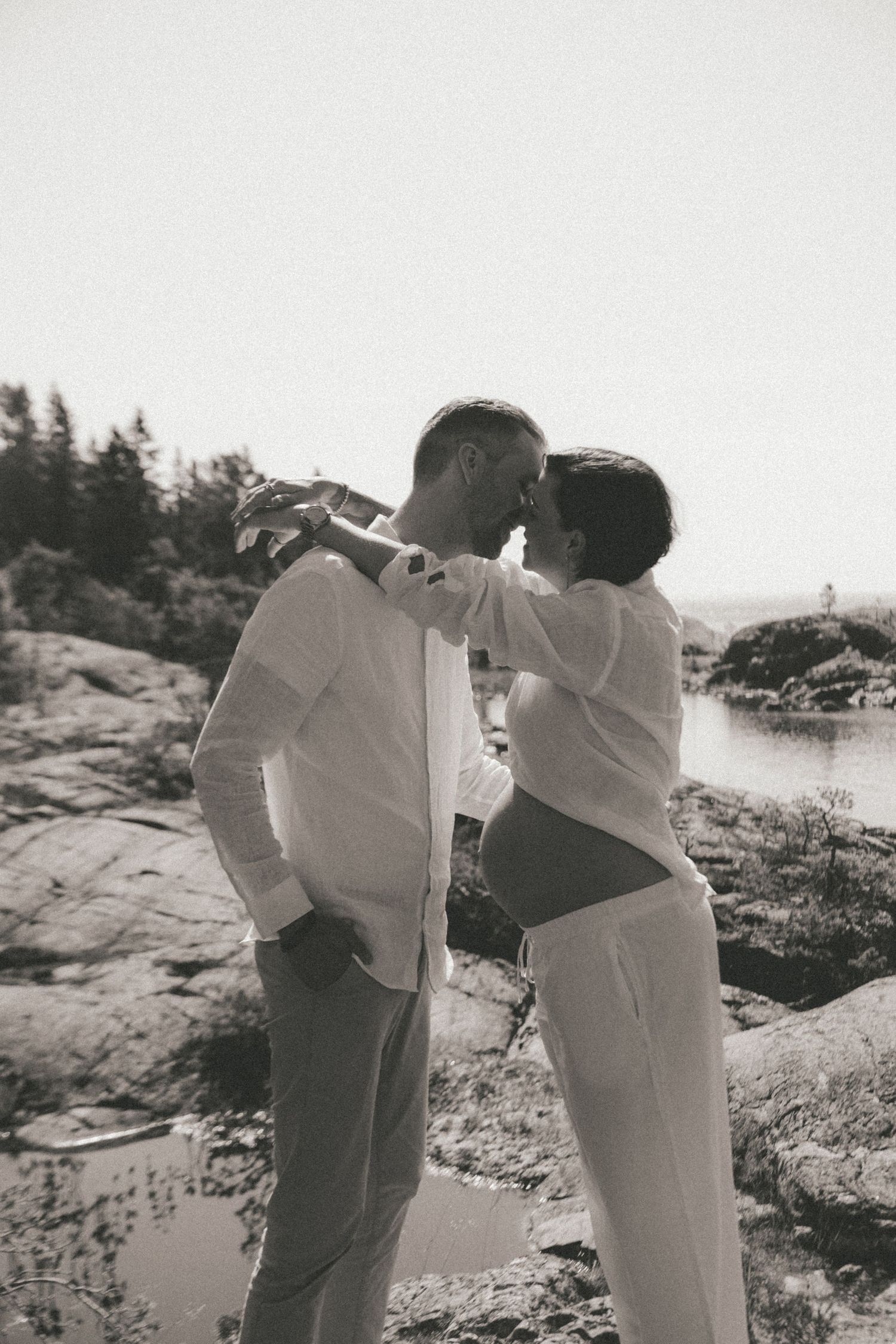A couple in white clothing shares romantic moments on rocky coastal terrain in black and white.