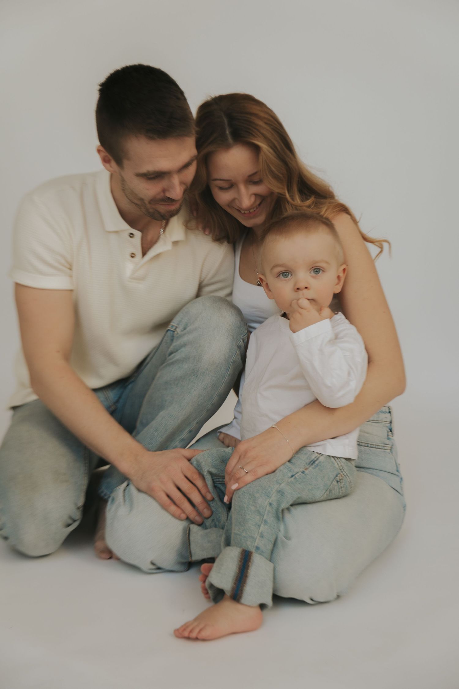 A family in casual attire sits close together on a white backdrop for an intimate studio portrait session.
