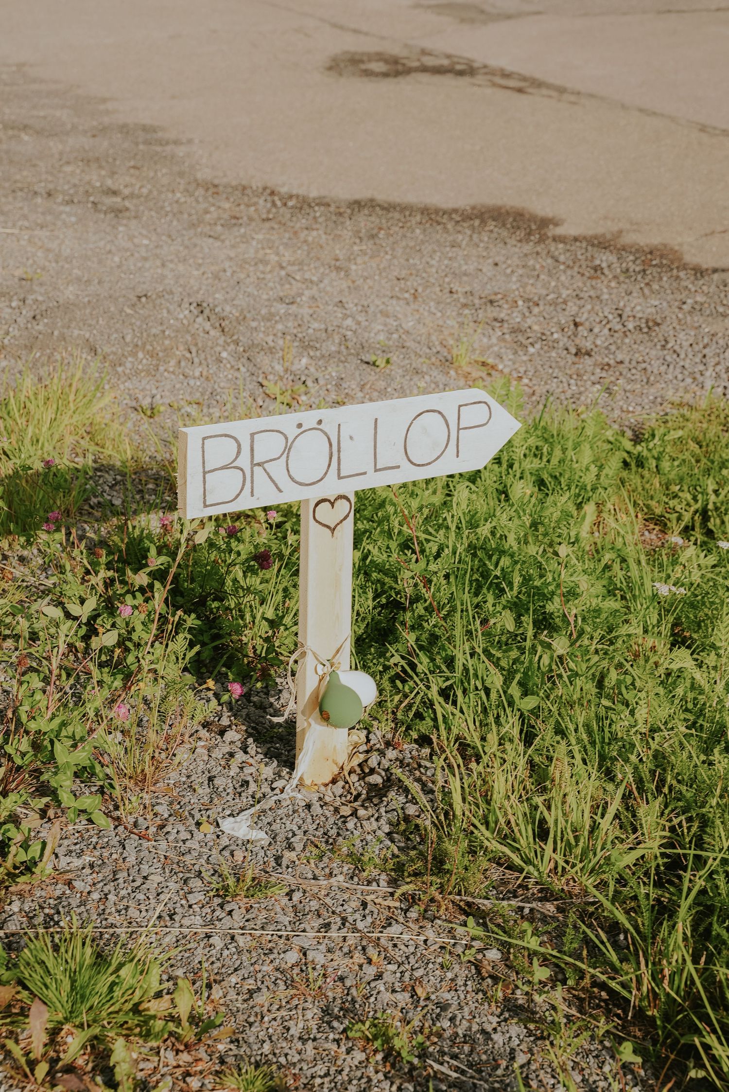 A white wooden cross marker with 'BROLLOP' text and a heart symbol stands in grassy gravel ground.