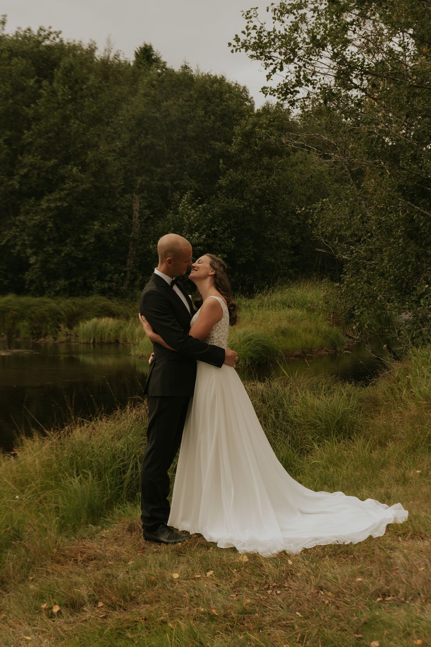 Wedding couple embraces near a scenic pond surrounded by lush greenery and trees.
