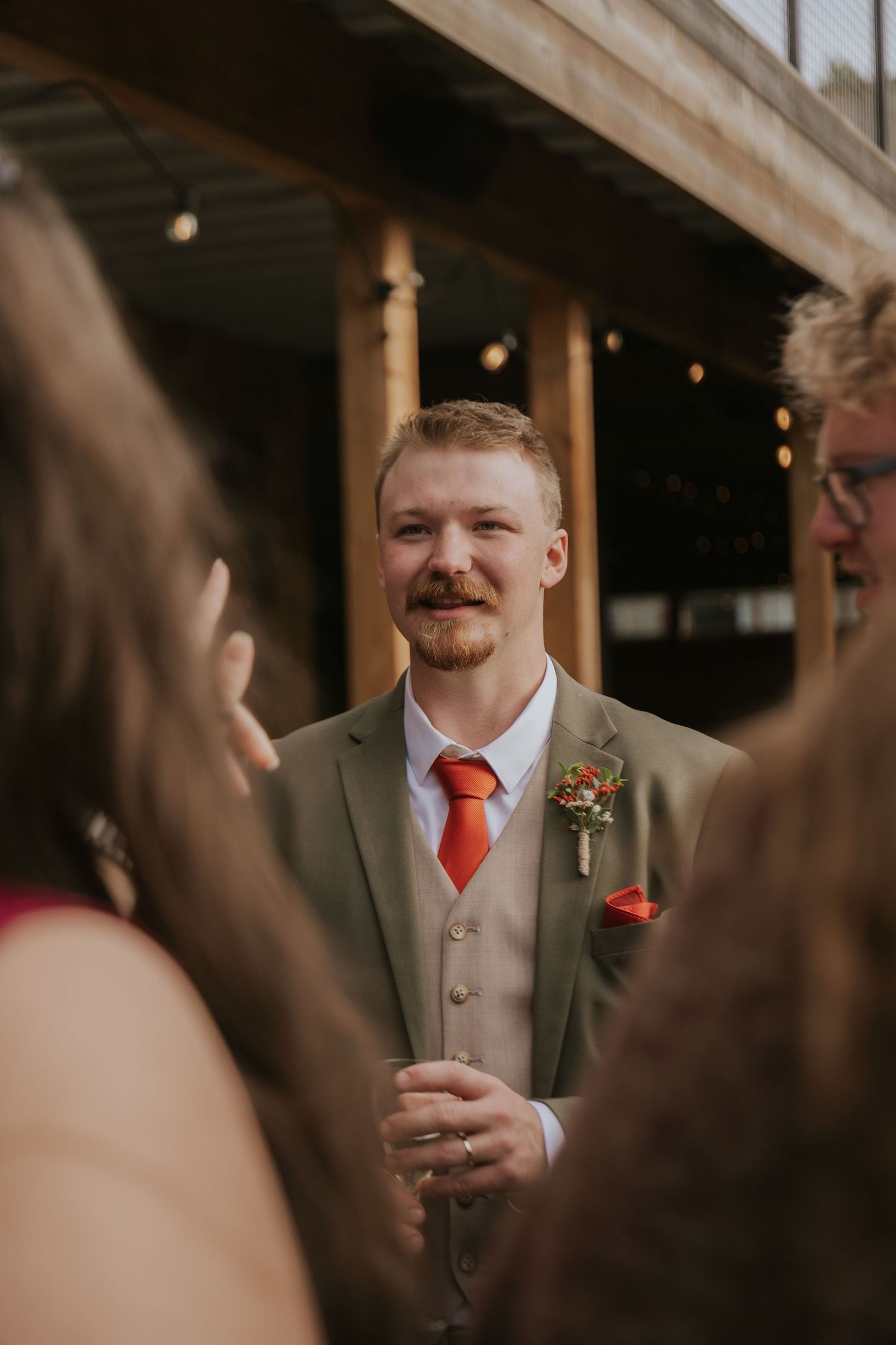 A guest in a tan suit and red tie mingles at an outdoor event with string lights overhead.
