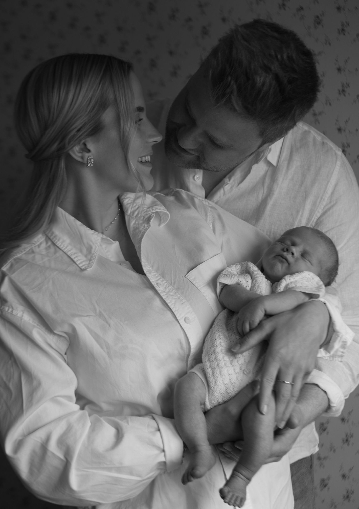 A tender black and white portrait of a couple in white shirts sharing a joyful moment with their newborn baby.