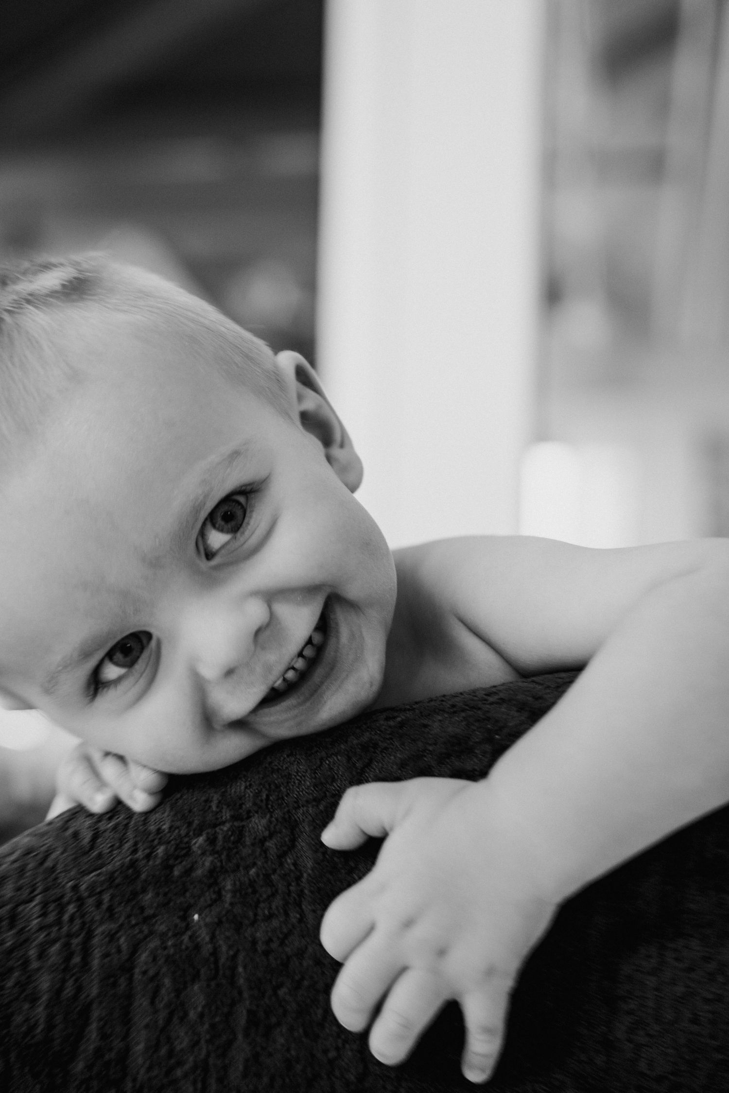 A black and white portrait of a joyful young child resting their head on a dark surface with a bright smile.