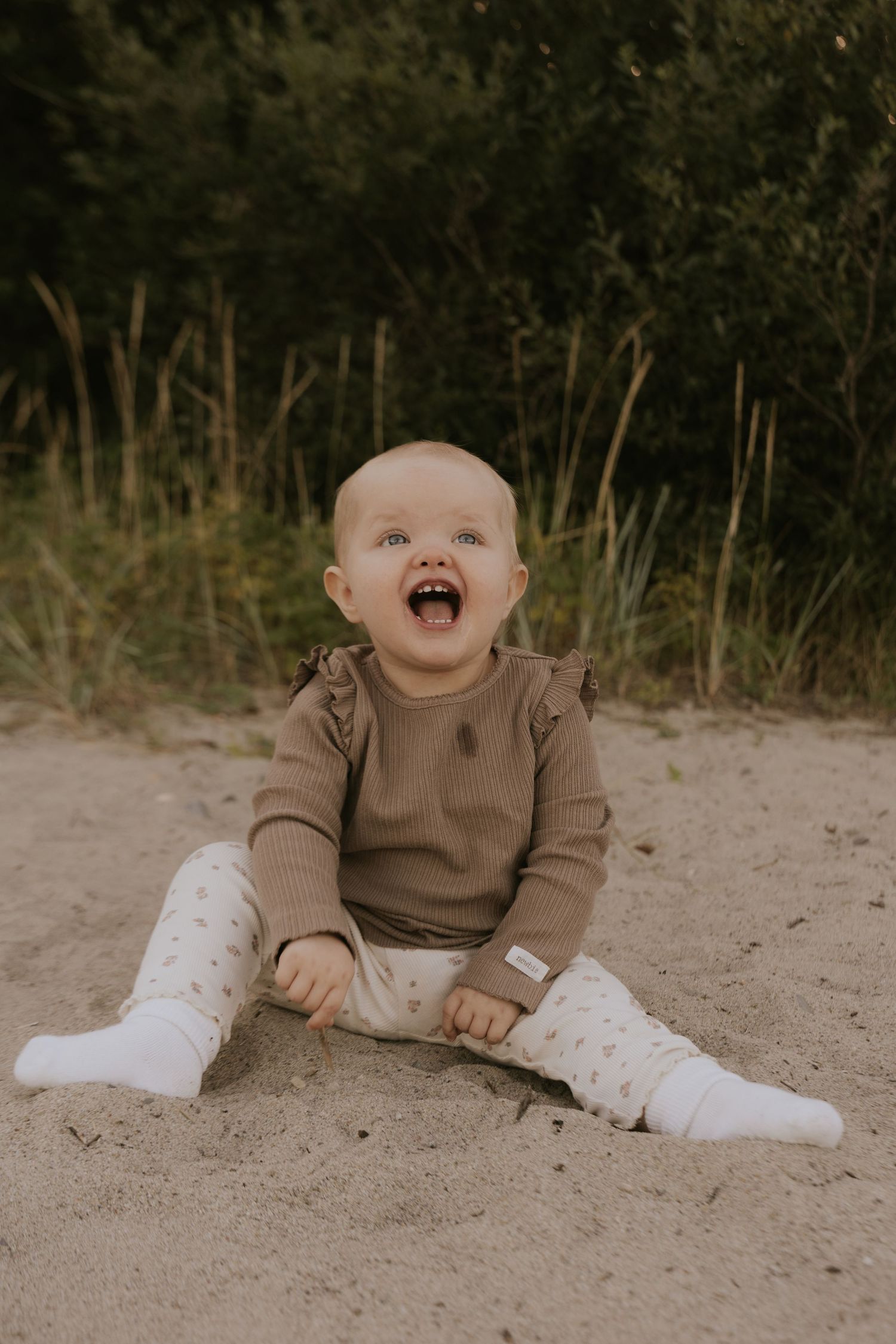 A laughing baby in a brown sweater sits on sandy ground with legs spread wide.