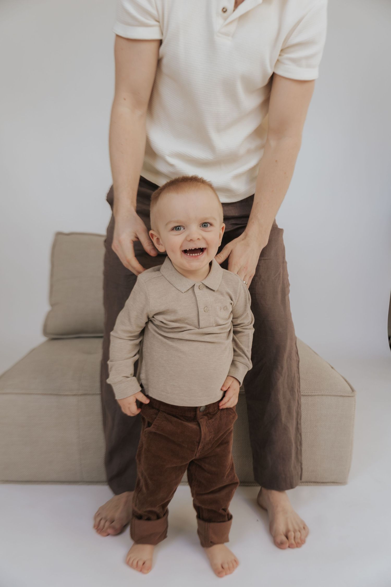 Baby in beige sweater and brown pants stands supported by parent in white shirt against neutral background.