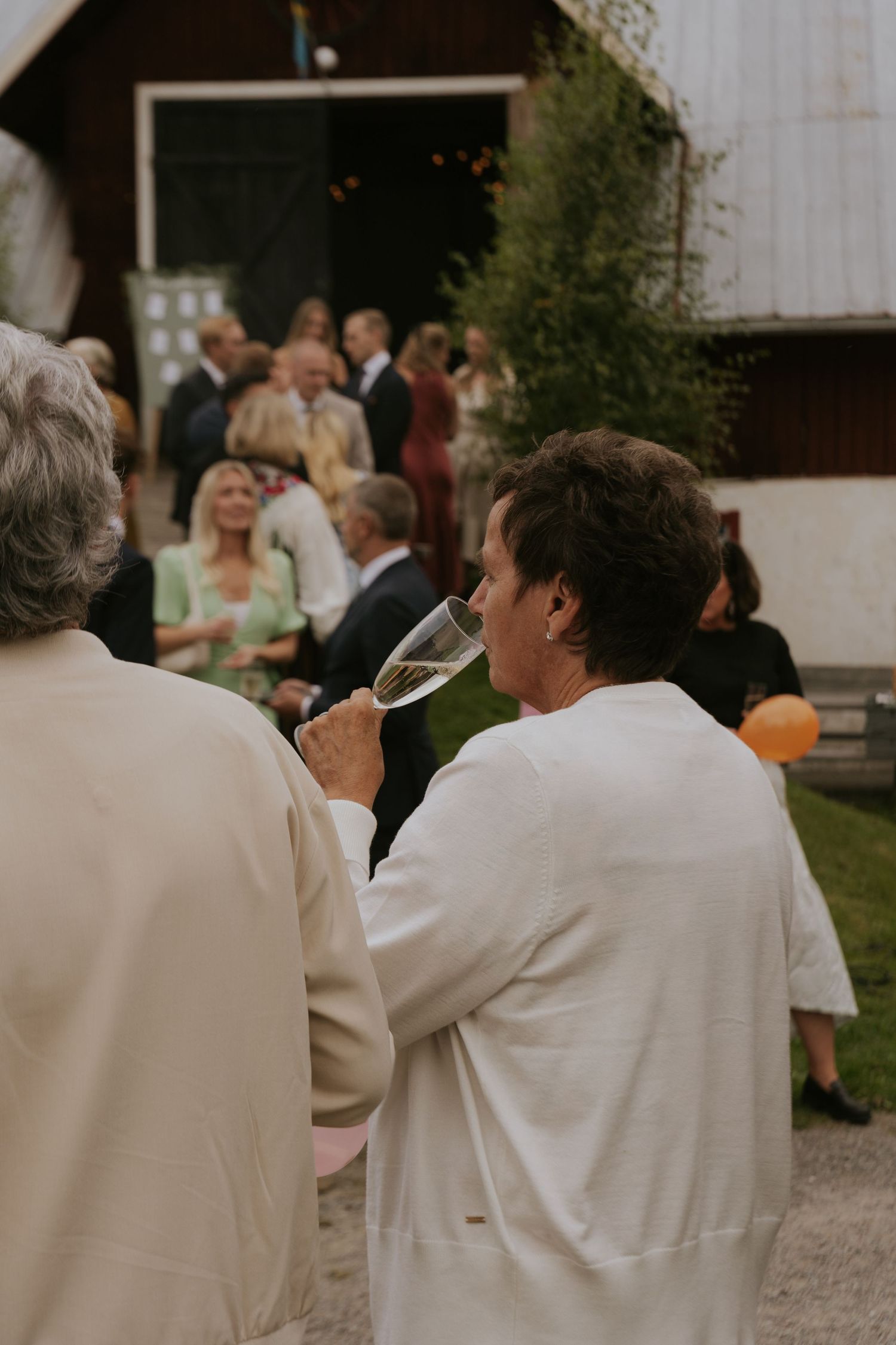 Wedding guests in white attire gathered outdoors at a rustic venue during a celebration.