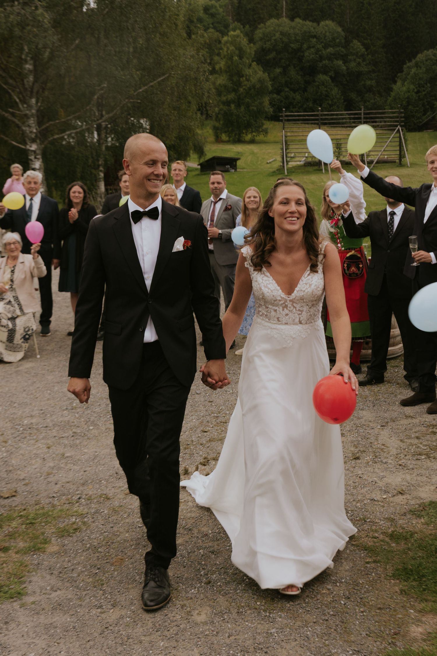 Wedding party walks together outdoors holding colorful balloons while celebrating on a gravel path.