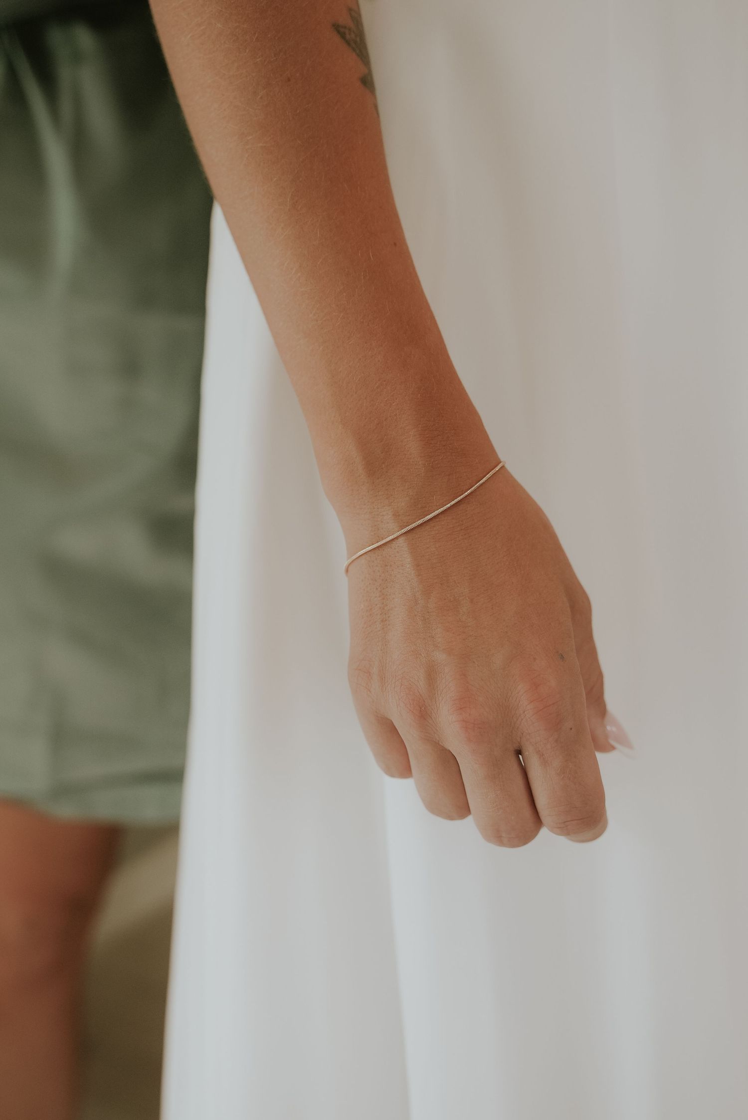 Close-up shot of a person's hand wearing a delicate gold bracelet against a soft white and green background.