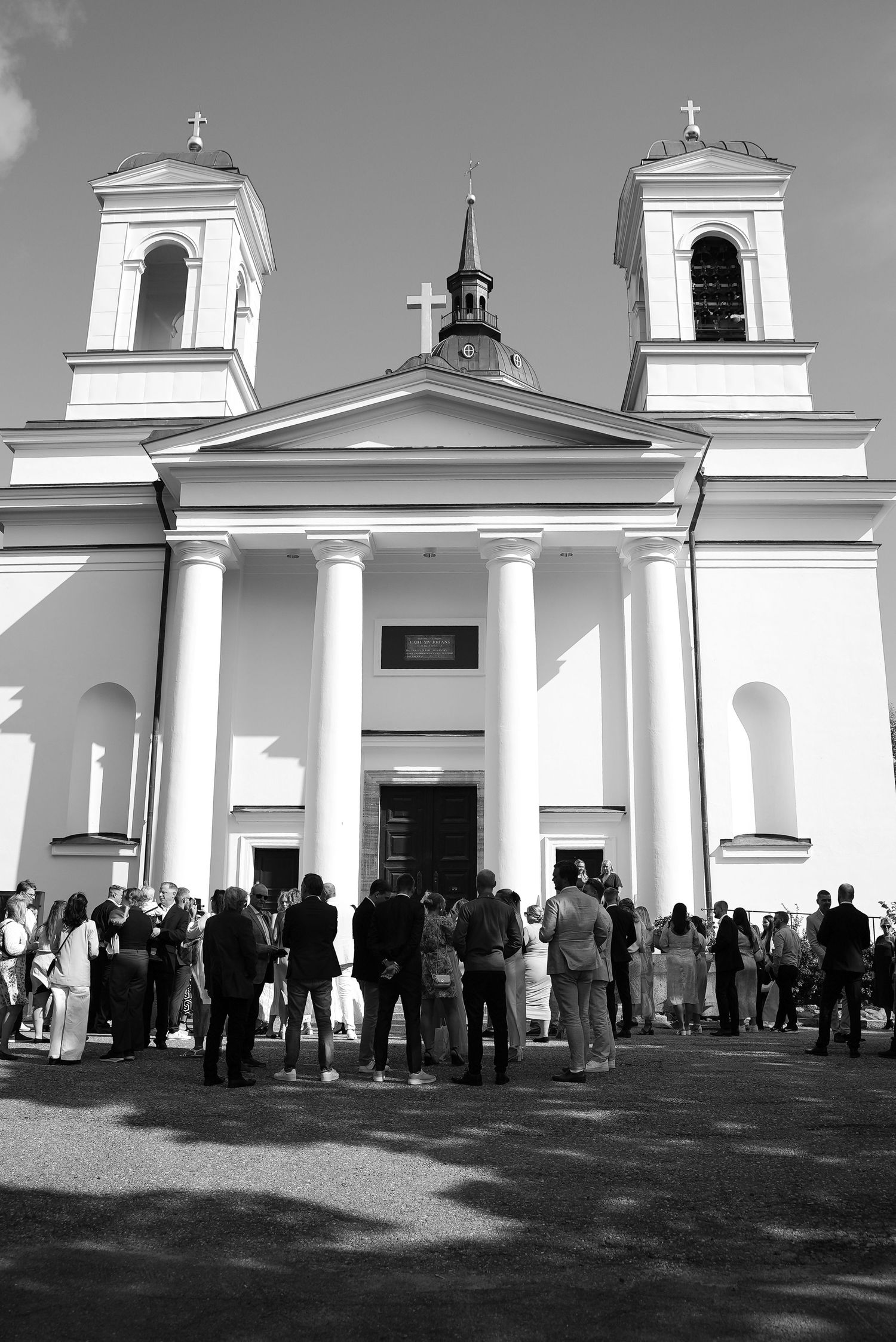 People gather outside a white colonial church with multiple steeples and arched windows during a daytime event.