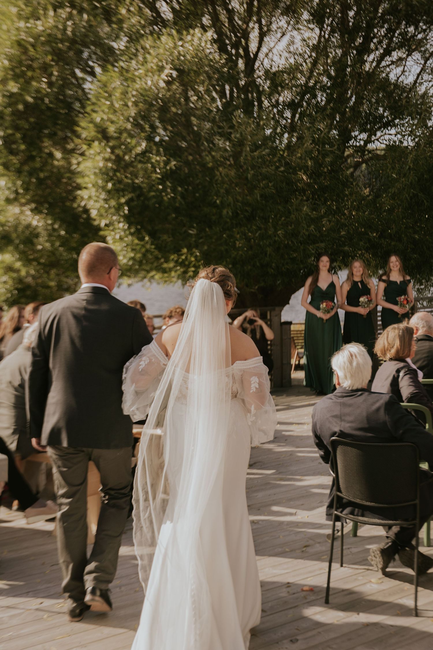 Outdoor wedding ceremony with guests seated on chairs and bride in flowing white dress.