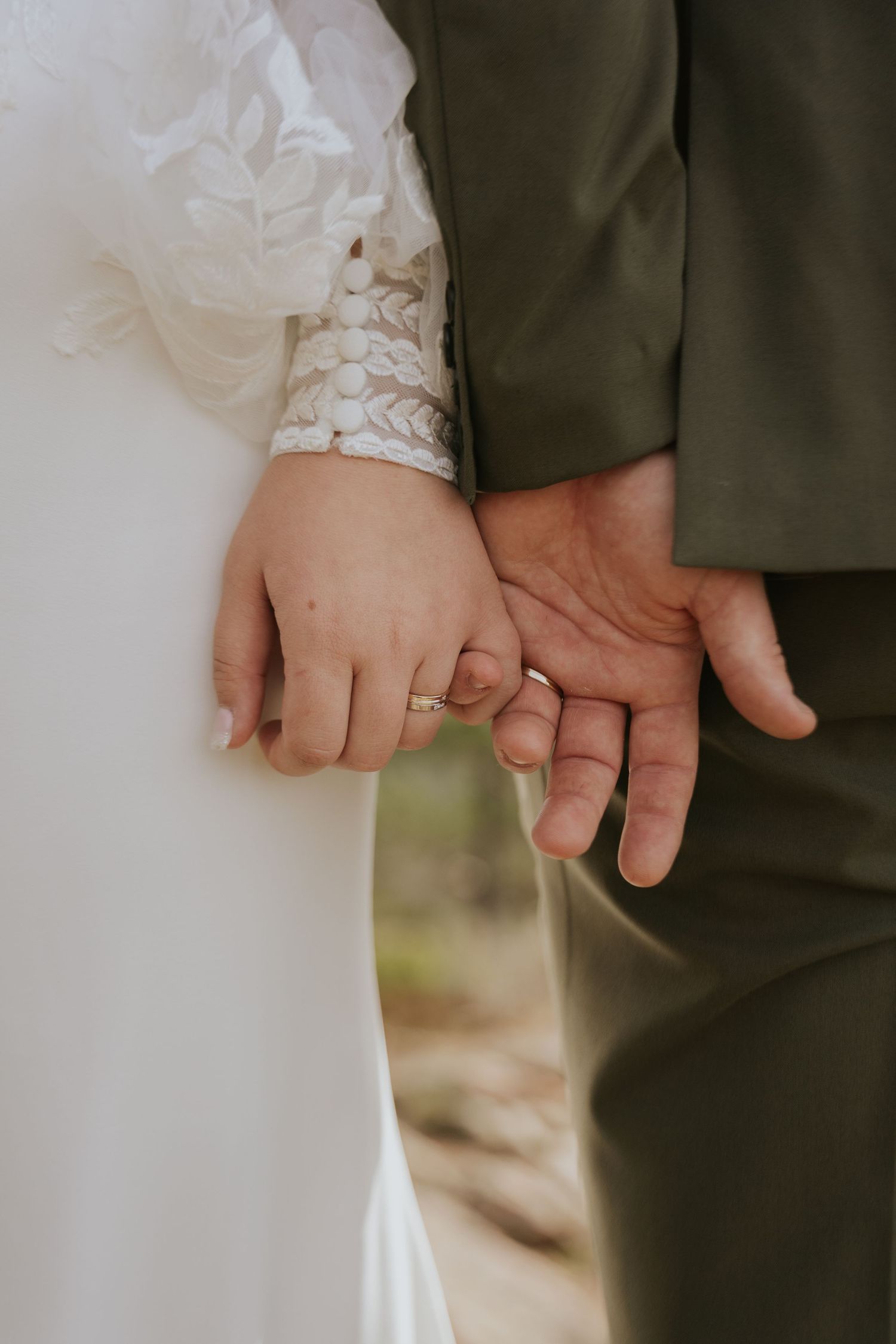 Close-up of wedding rings and joined hands against a white dress and dark suit background.