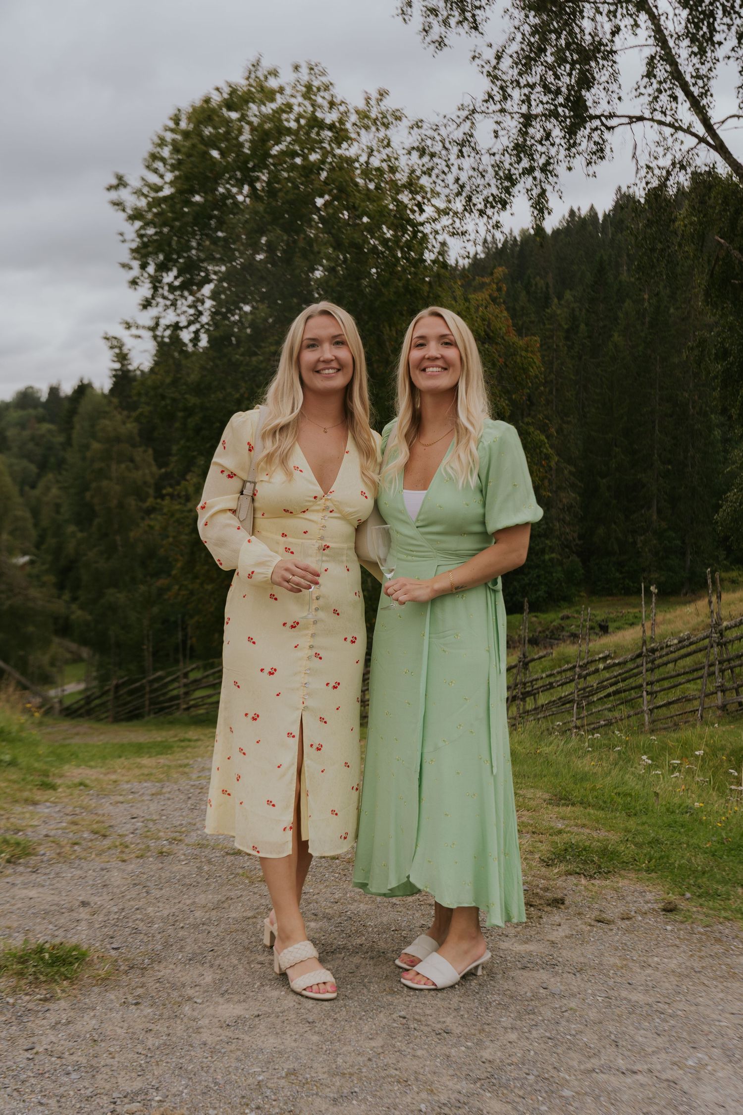 Two people in coordinating pastel dresses pose together on a gravel path with green hills and trees in the background.