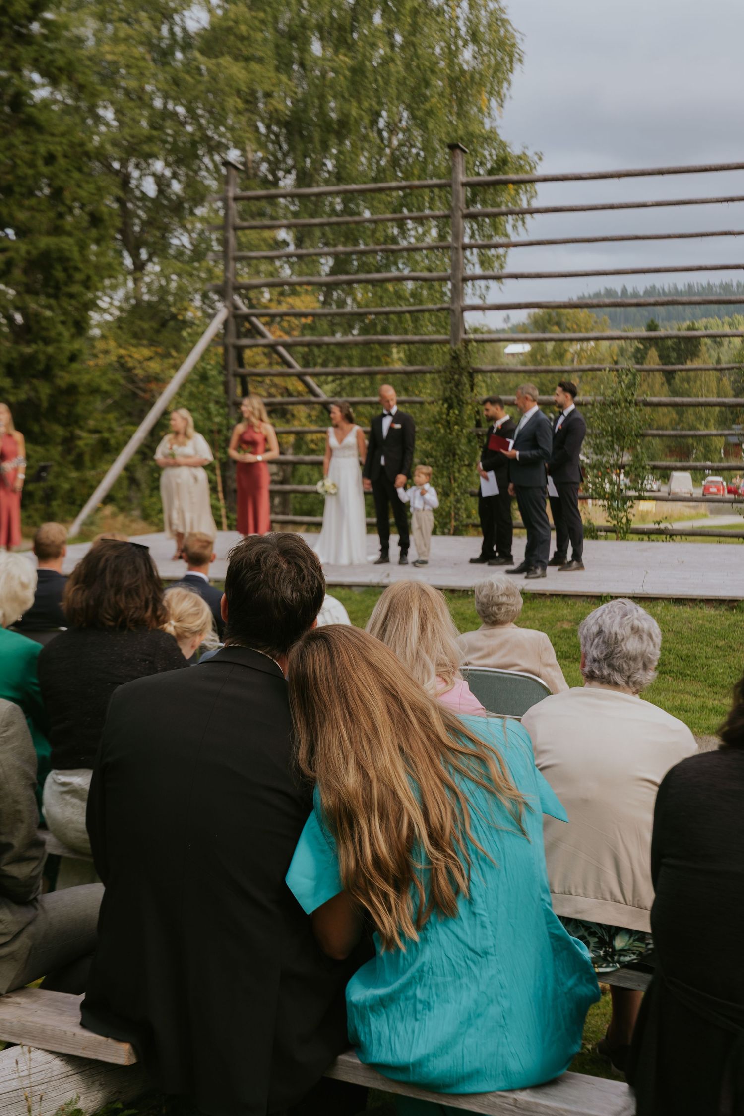 Wedding guests seated outdoors watching a ceremony on an elevated platform with wooden railings.