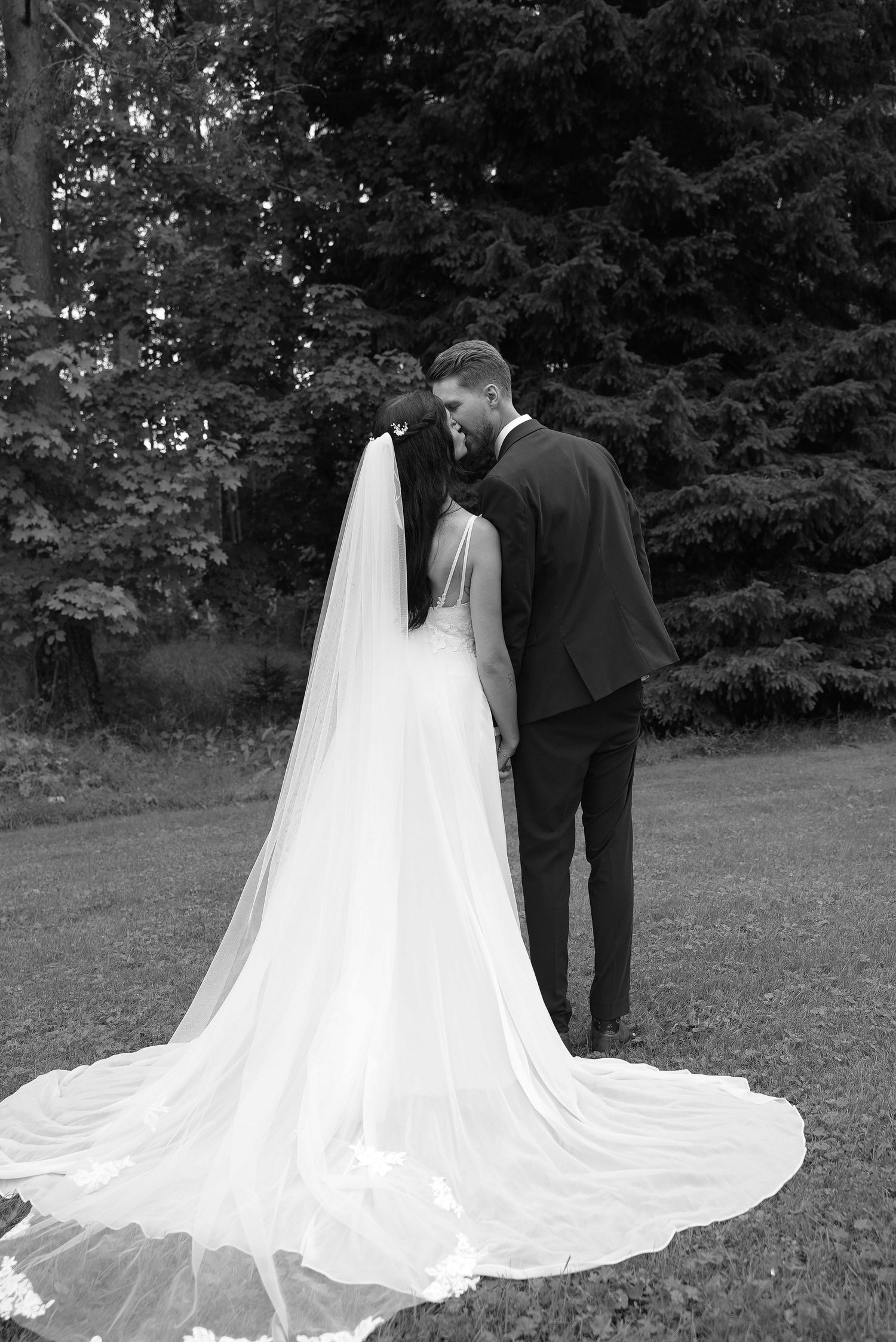 A romantic black and white wedding photo showing a couple in formal attire with a flowing white veil against a wooded backdrop.