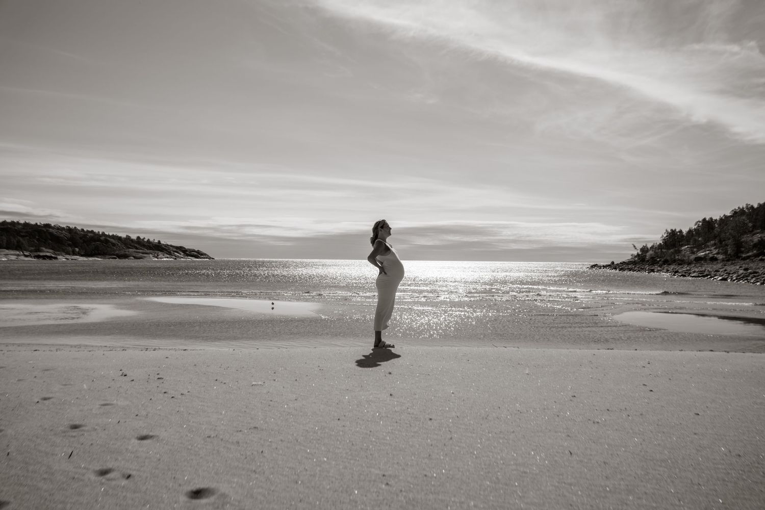 A silhouetted figure stands on an empty beach at sunset with sparkling water and hills in the background.