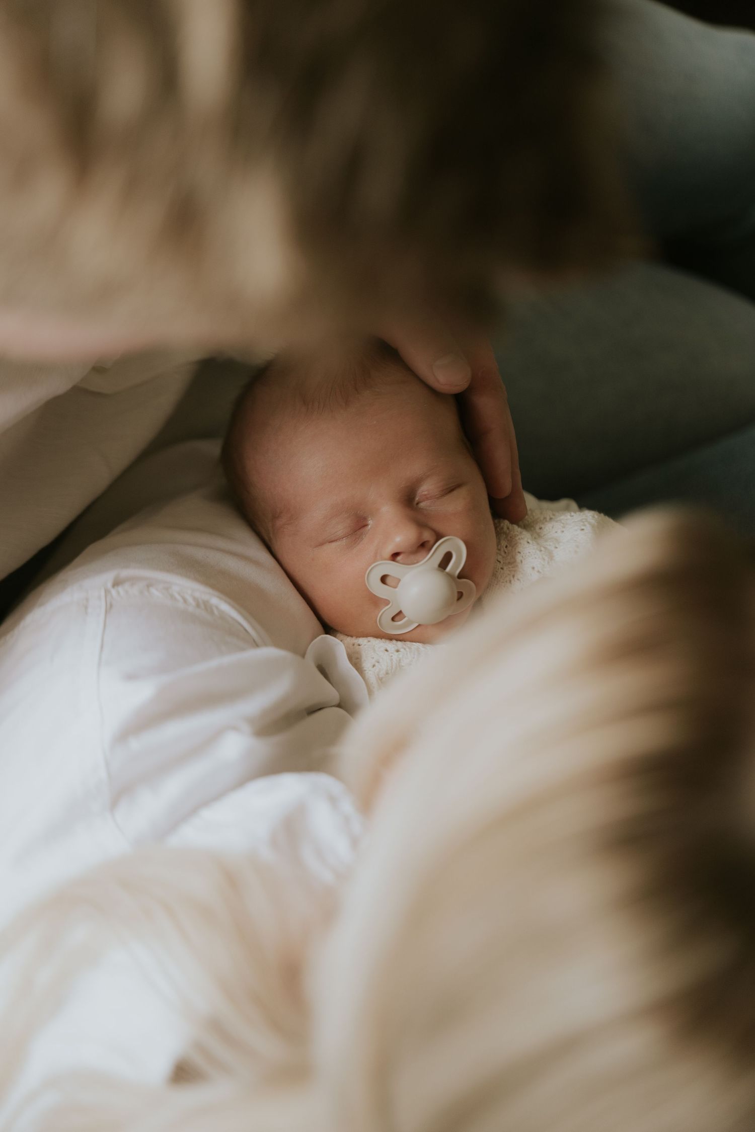 A sleeping newborn baby swaddled in soft white blankets rests peacefully in a dimly lit setting.