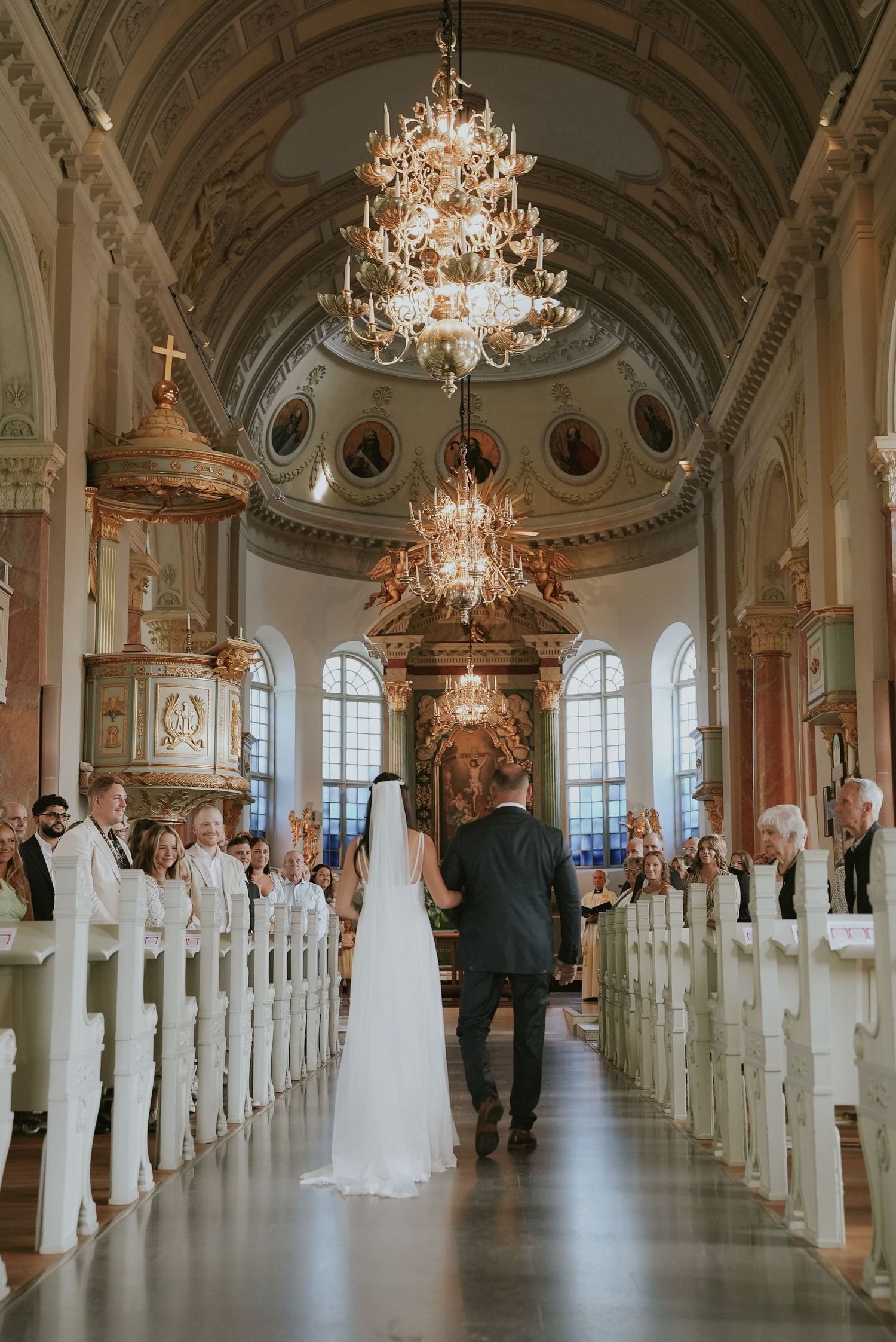 Elegant wedding ceremony venue with ornate chandeliers and white chairs in a grand ballroom with arched ceilings.