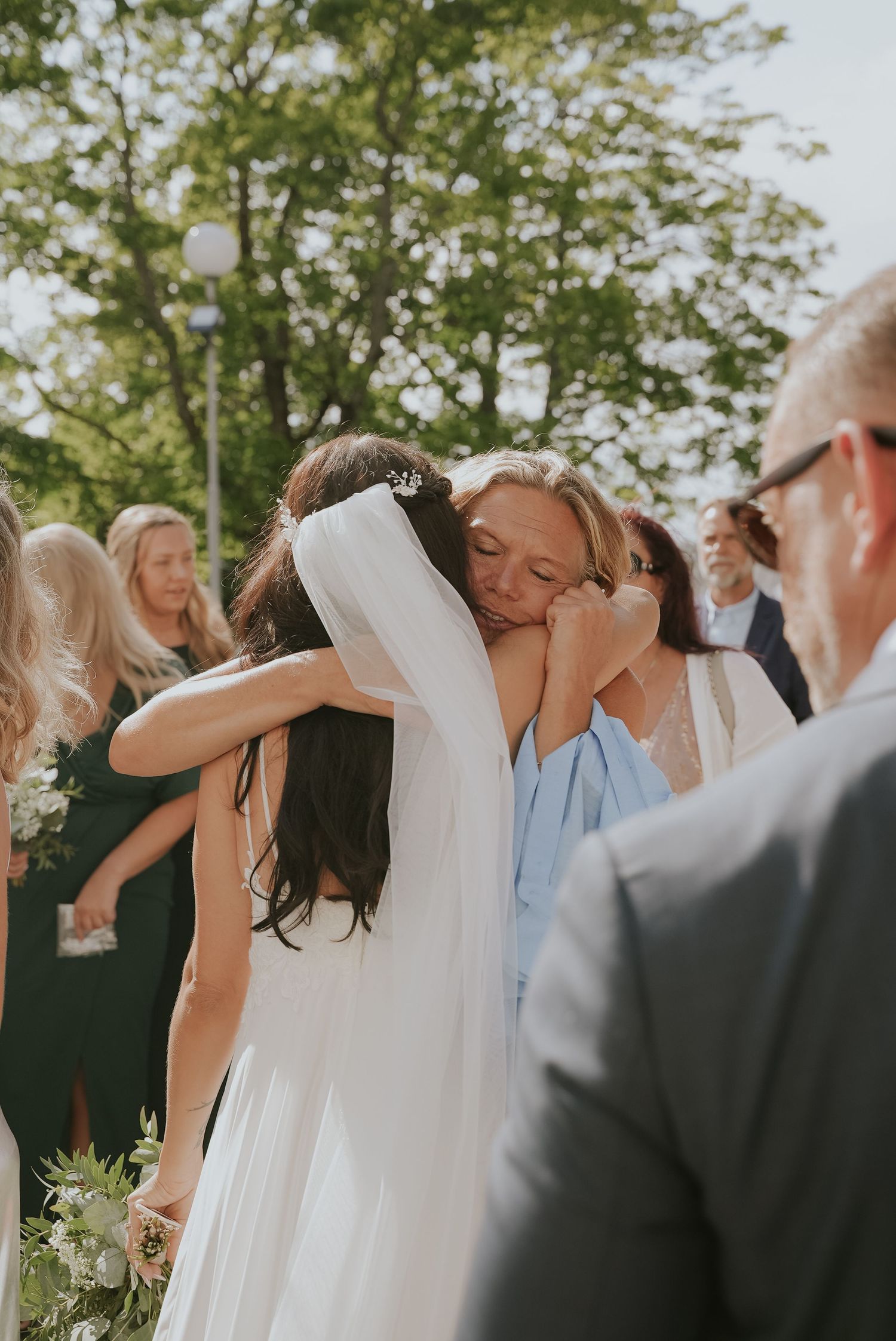 Wedding guests share emotional embraces during an outdoor ceremony beneath green trees in natural sunlight.
