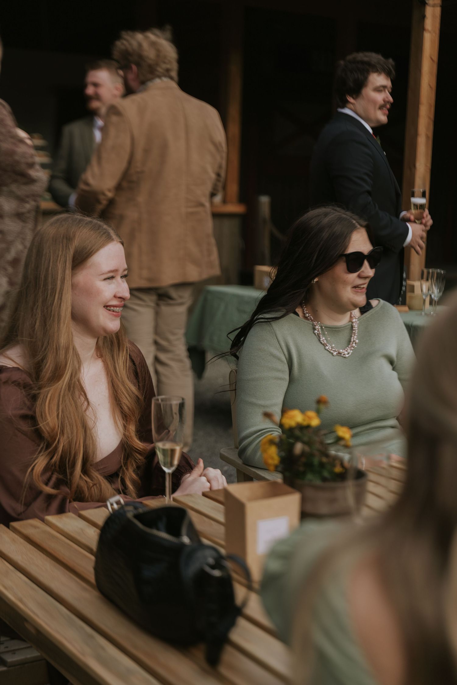 Two friends enjoying drinks at an outdoor wooden table with yellow flowers and sunglasses on a casual afternoon.