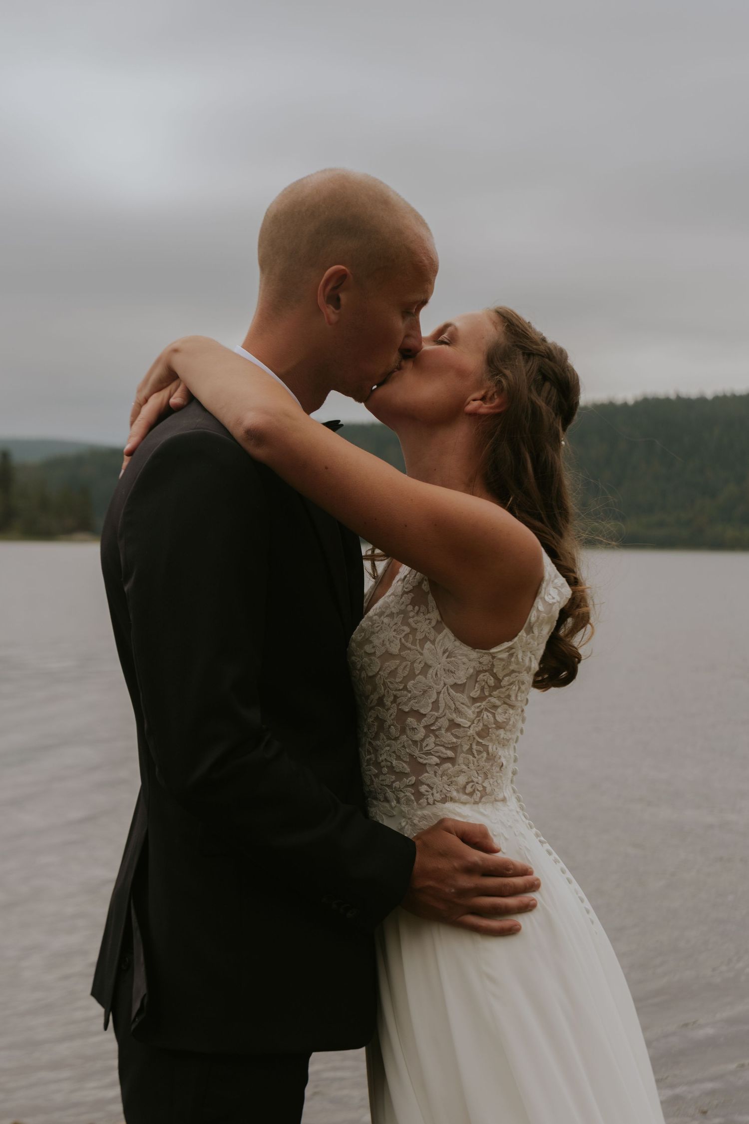 Couple in wedding attire embracing passionately by lakeside.