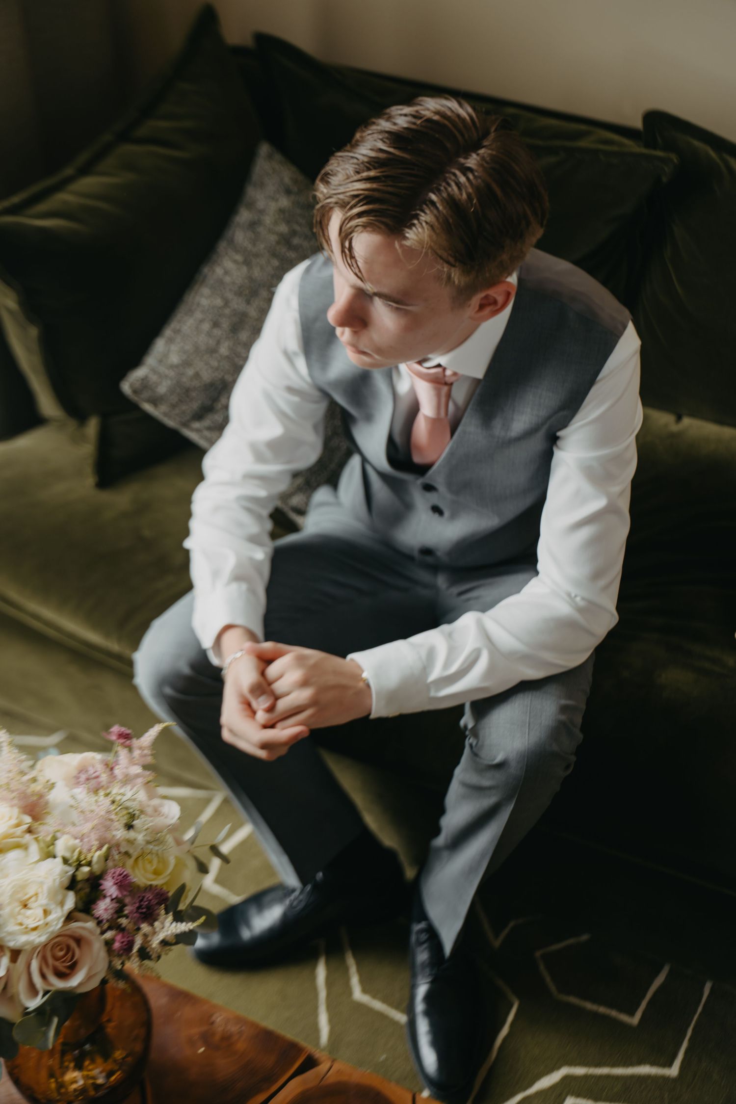 Person in formal attire sitting on green velvet couch near floral arrangement.