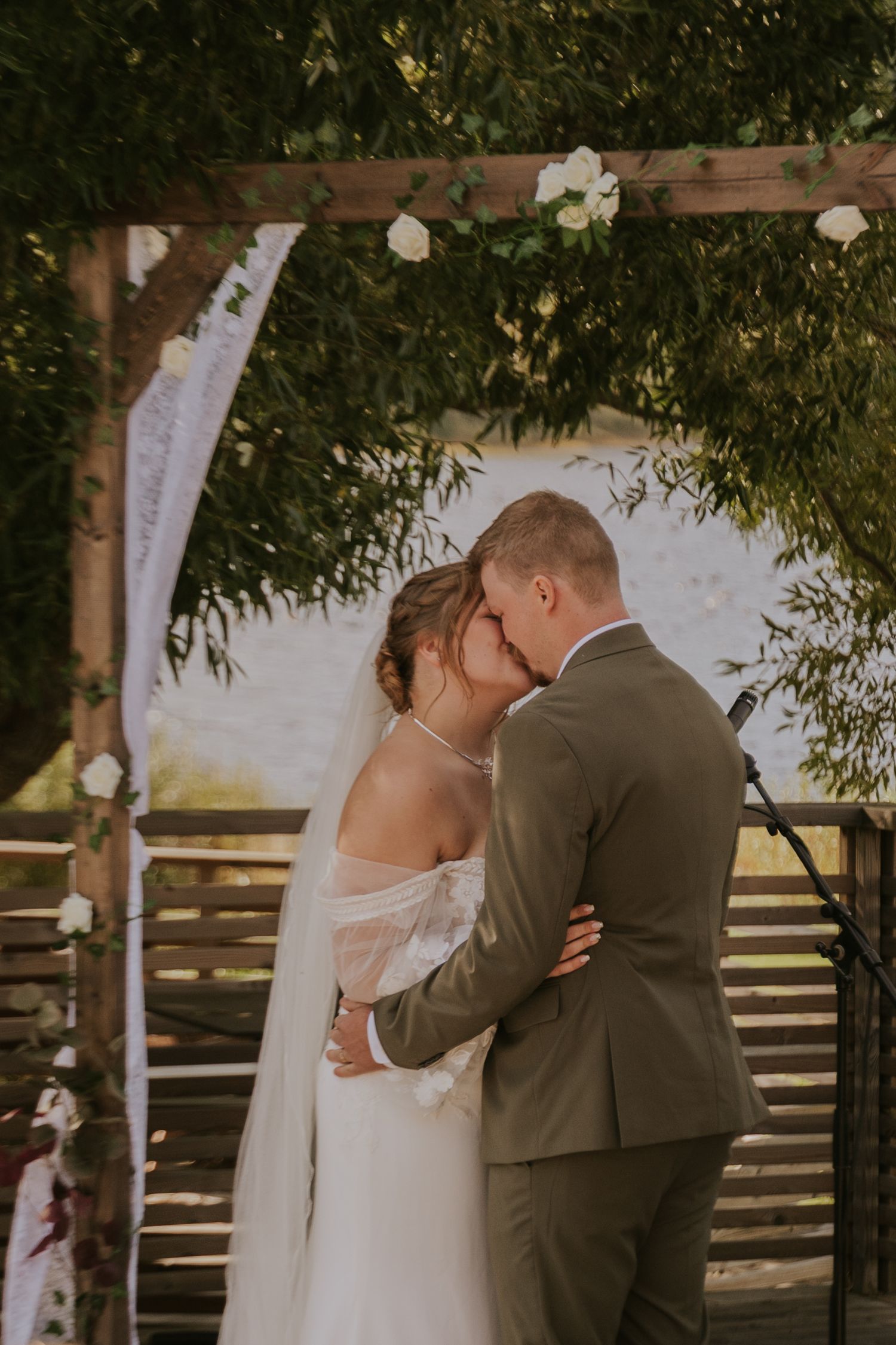 Intimate moment between newlyweds during outdoor wedding ceremony under wooden pergola.