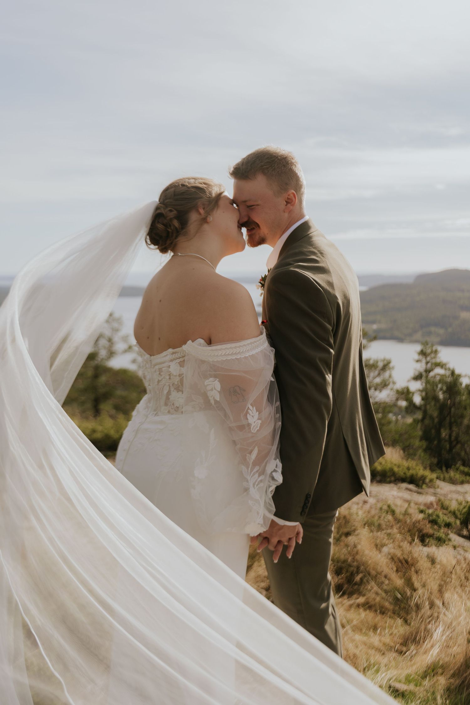 Bride's flowing veil catches the wind as couple shares romantic moment on clifftop.