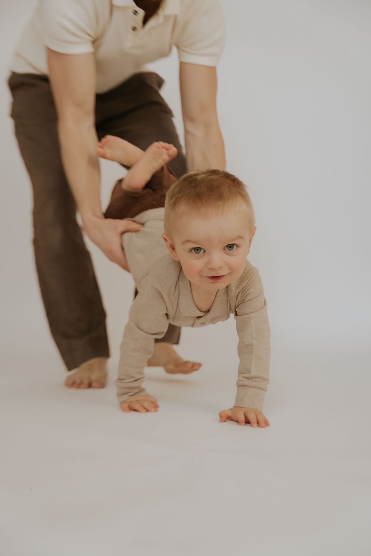 A baby in beige clothing crawls and explores on a white studio floor while supported by adult hands.