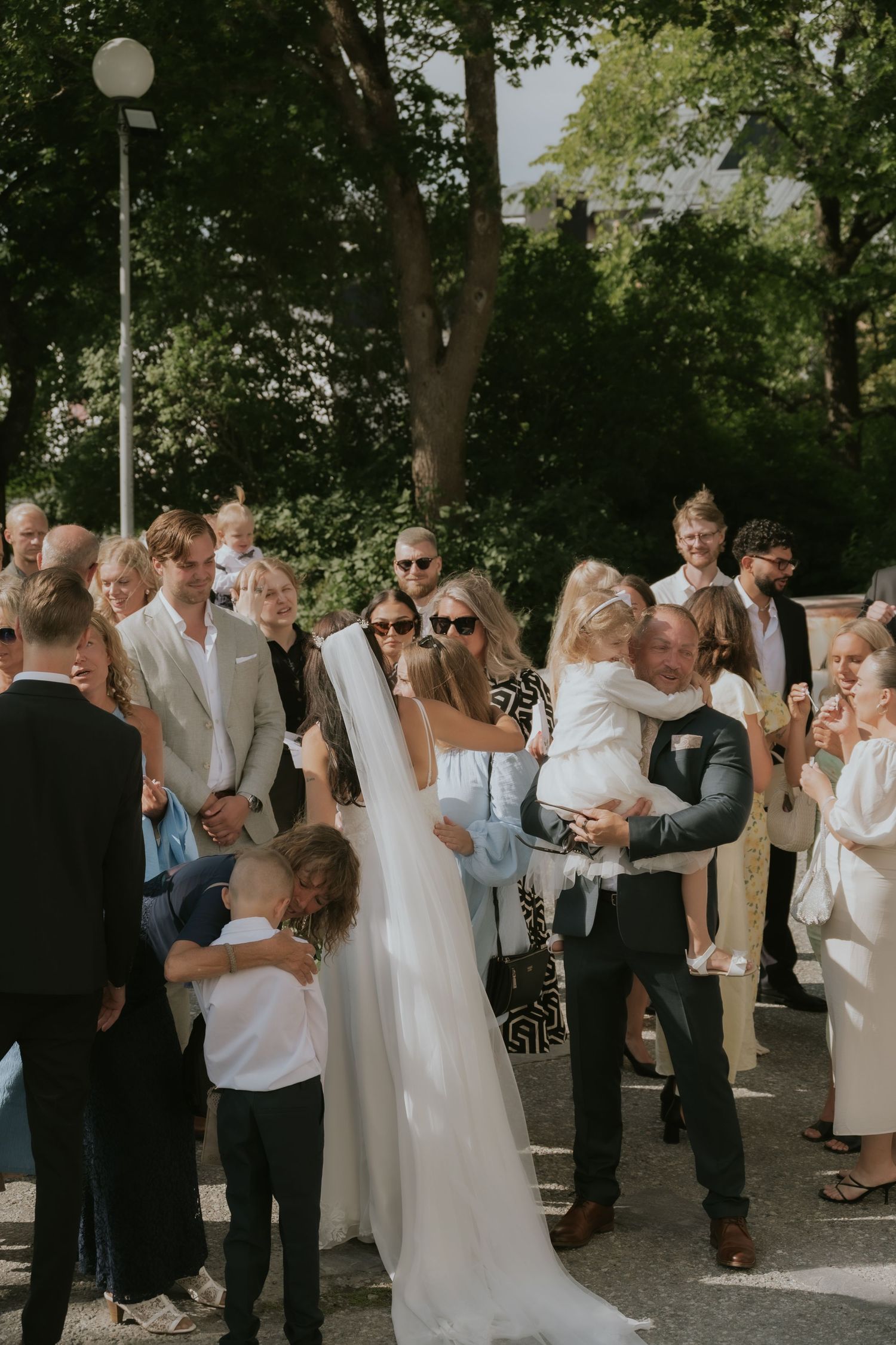 Wedding guests gather outdoors under trees during a summer celebration in white and casual attire.