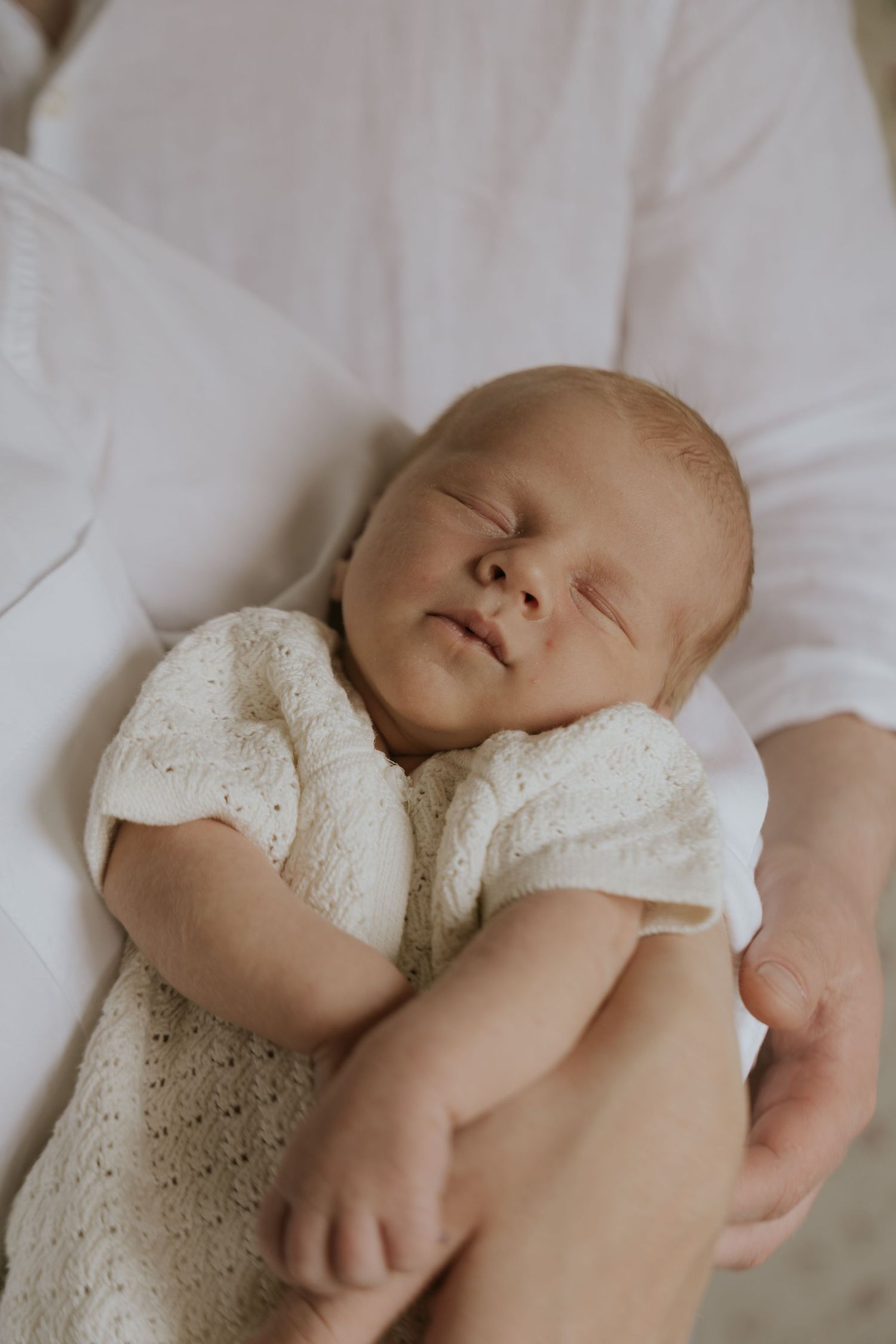 A sleeping newborn baby wearing a white lace dress lies peacefully on soft white bedding.