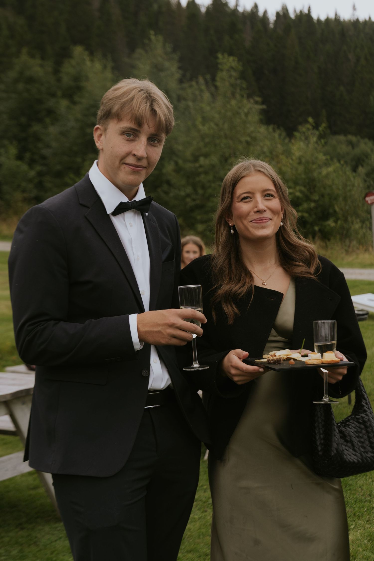 A couple in formal attire share drinks and appetizers at an outdoor event with forest scenery in the background.