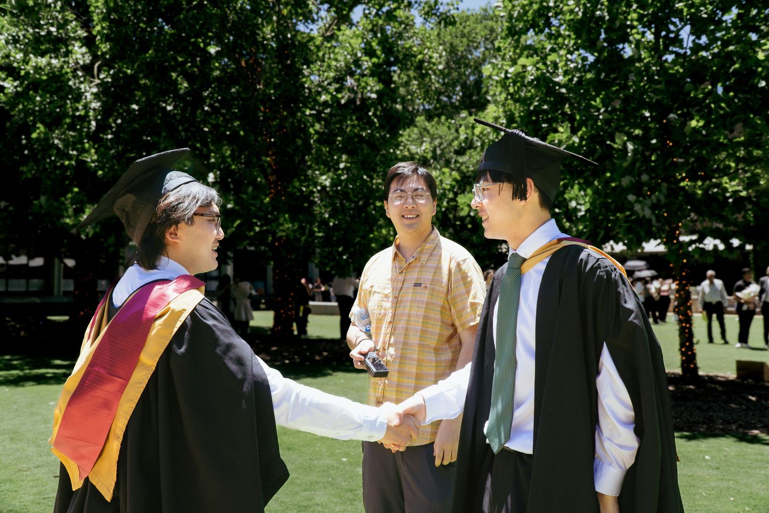 Two men shaking hands with another man looking at the camera at the Library Lawn at University of New South Wales.