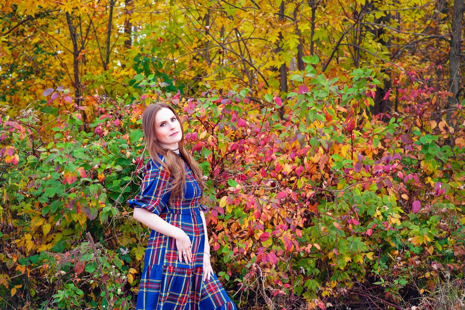 Young woman in blue plaid dress posing among vibrant red and yellow autumn foliage.