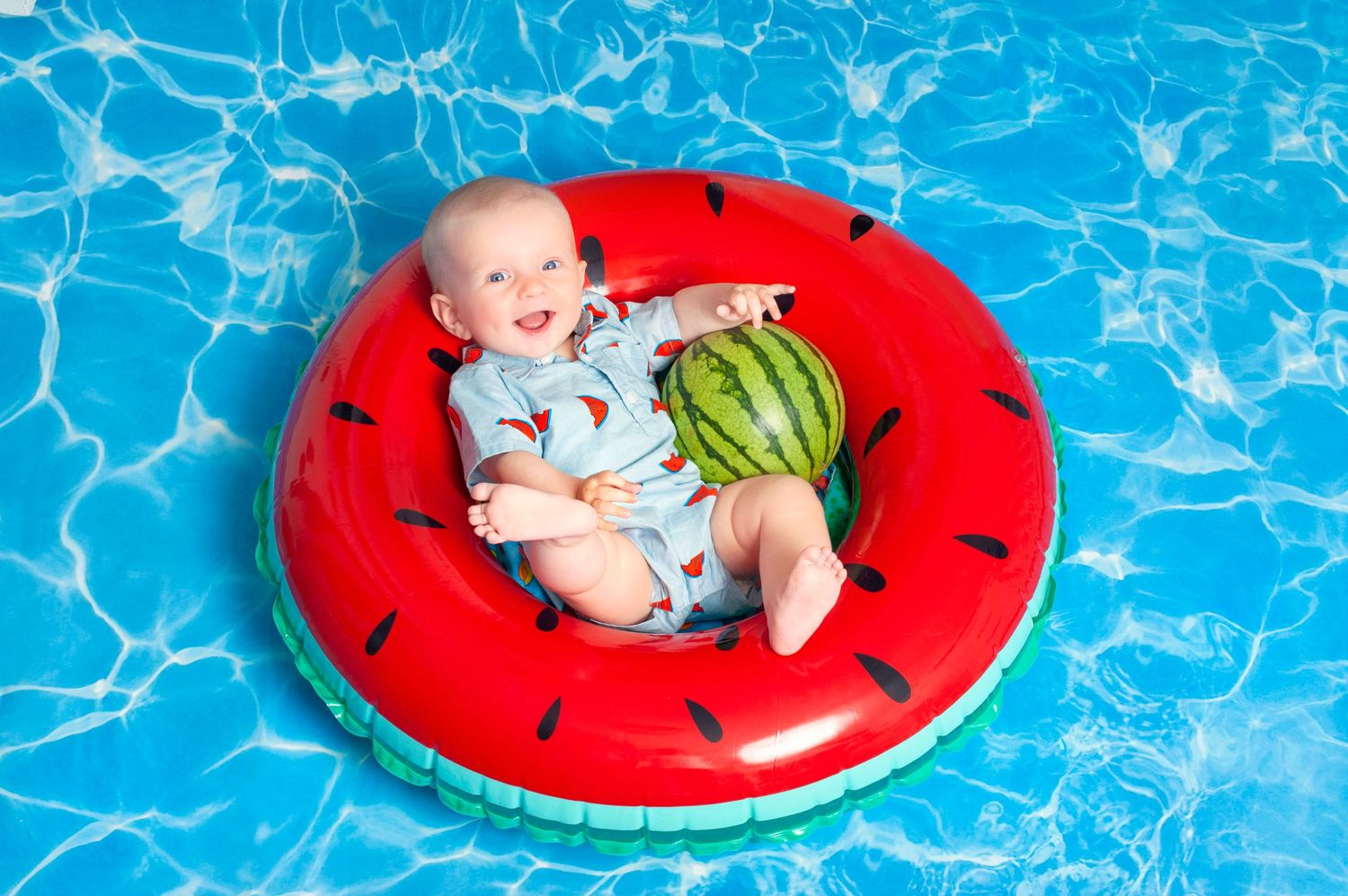 Happy baby holding a watermelon in a watermelon-themed inflatable ring in a blue pool.