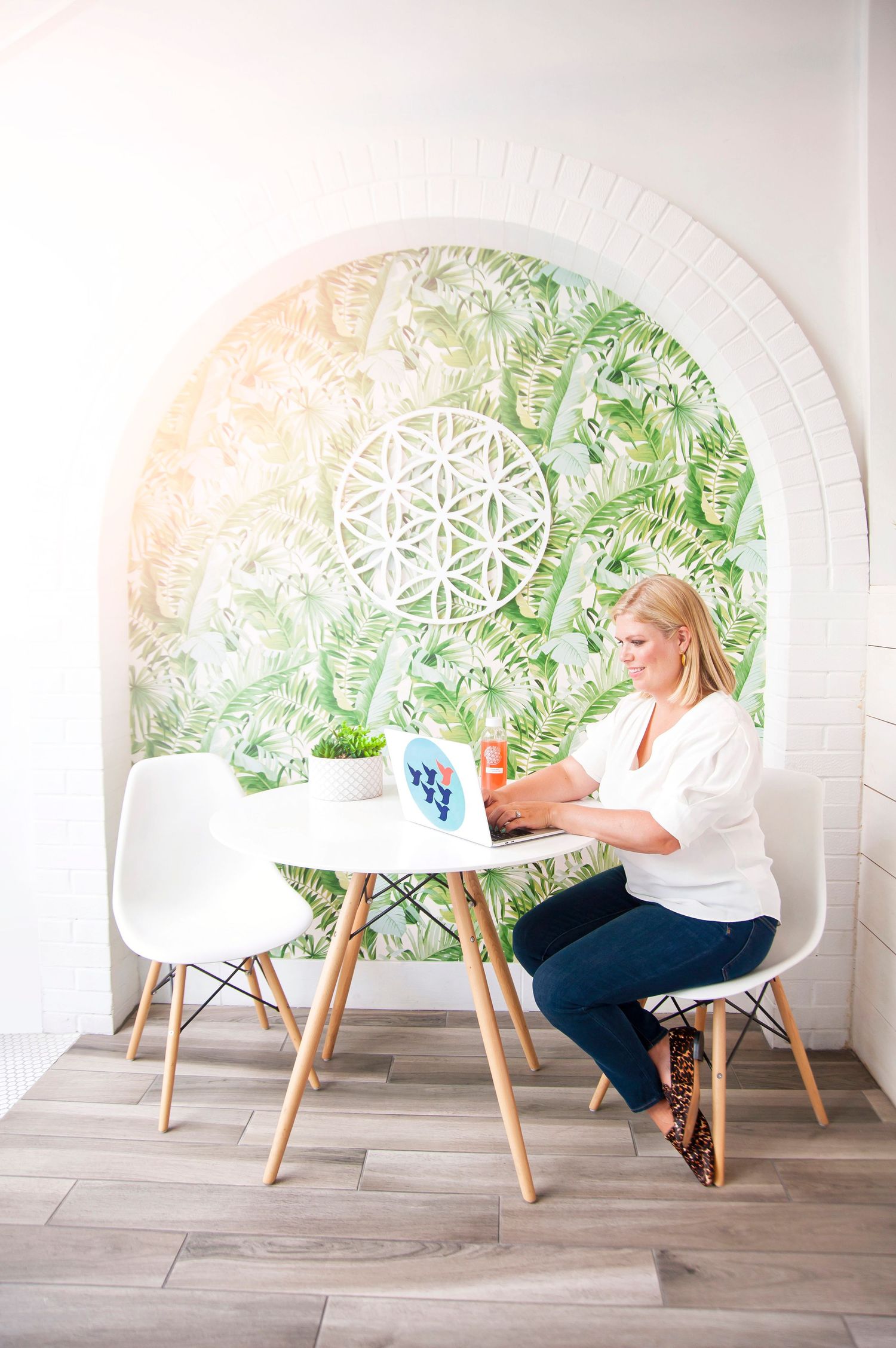 Blonde woman working on laptop at white table in bright room with tropical arch mural.