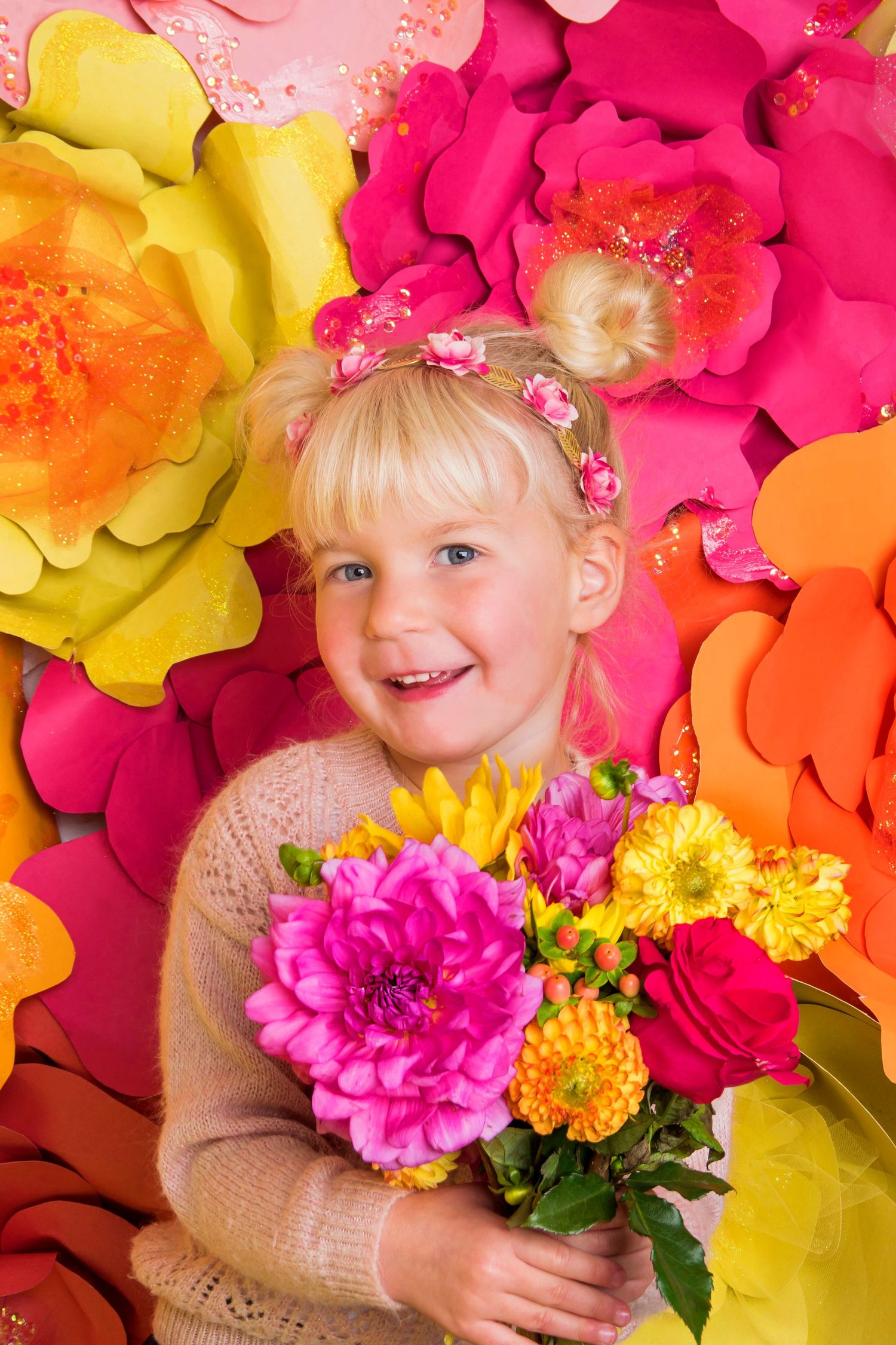 Smiling blonde girl holding colorful bouquet of pink and yellow flowers against vibrant paper flower backdrop.