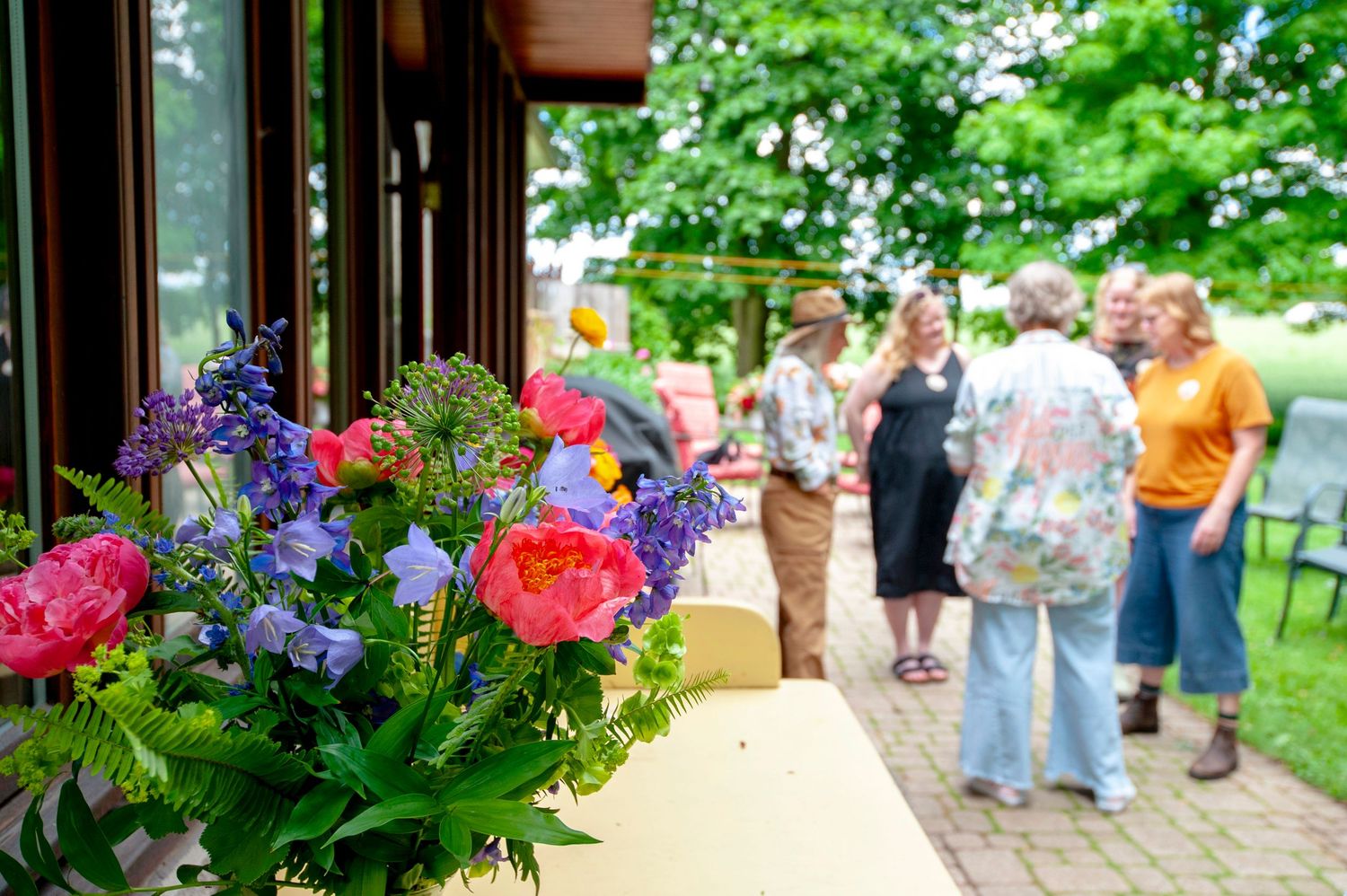 Vibrant pink peony and blue flower arrangement in foreground with guests socializing on a garden patio.