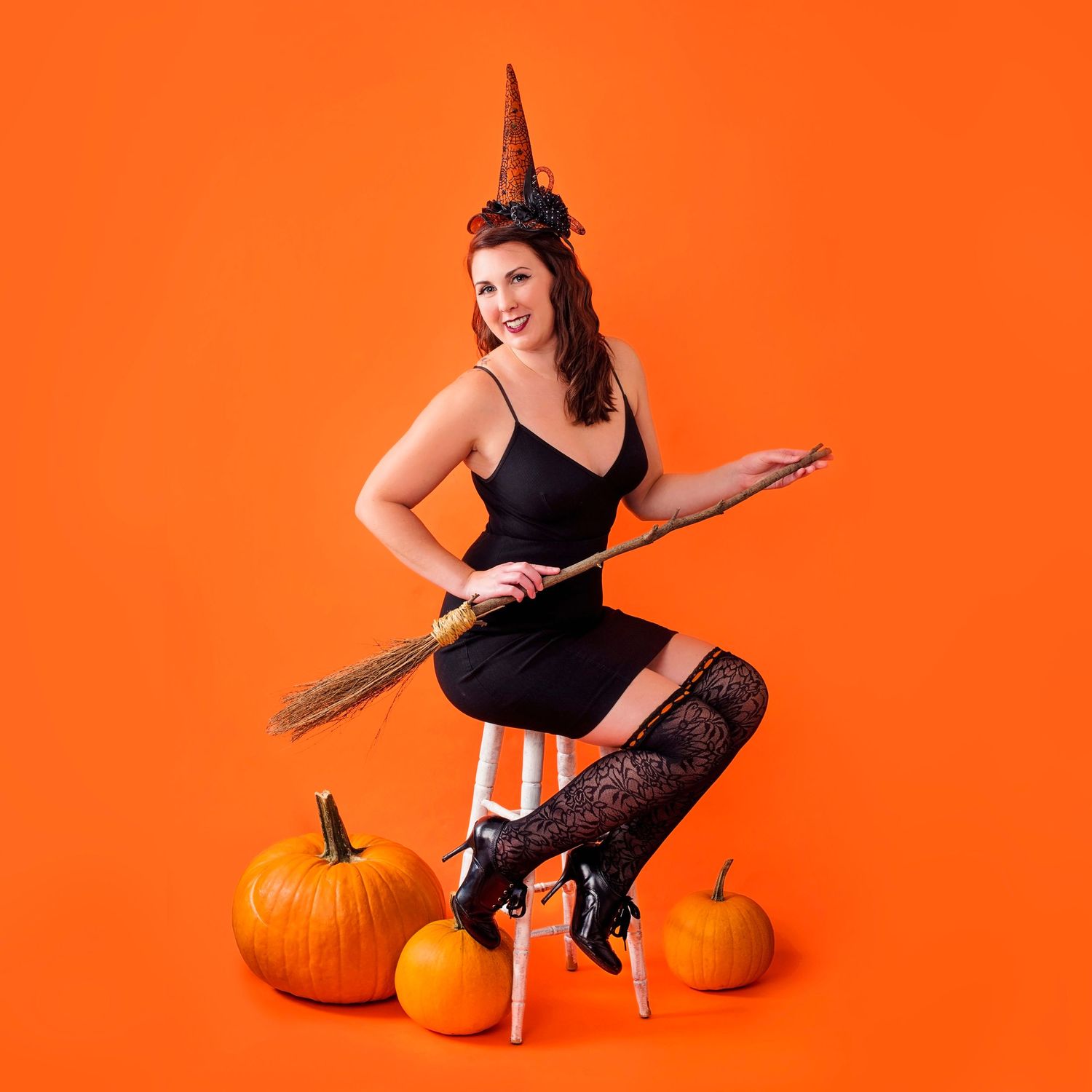 Woman in witch costume with broom and pumpkins poses on stool against orange background for Halloween.