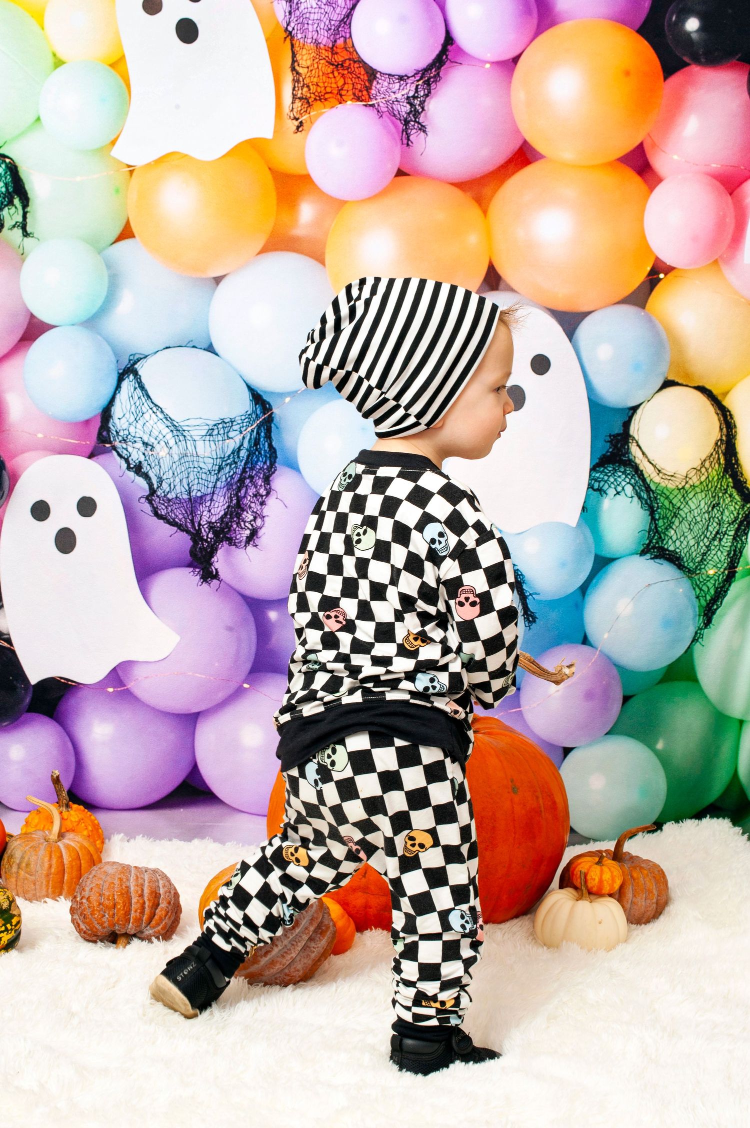 Young child in checkered Halloween outfit walking near pumpkins and colorful balloon backdrop.
