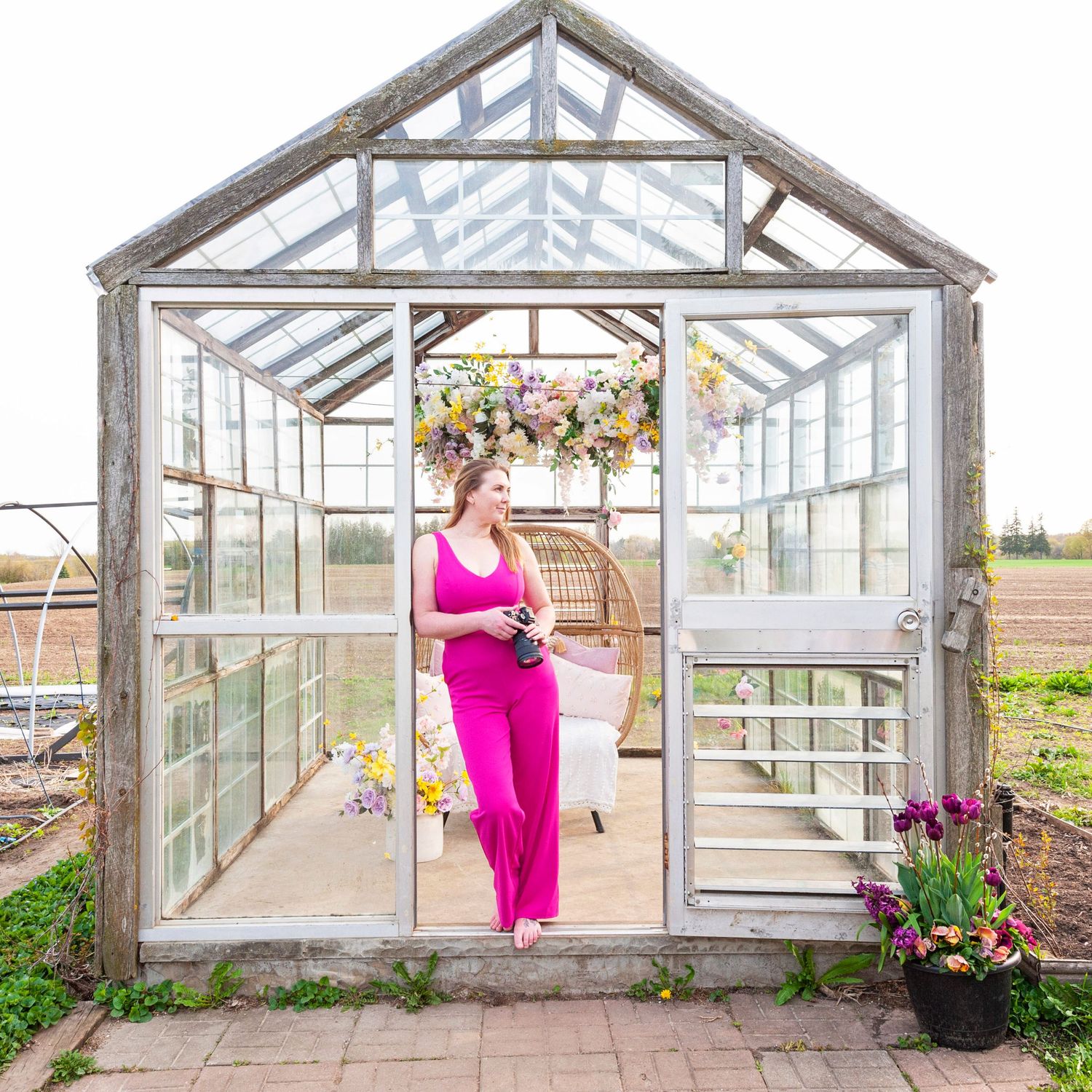Woman in a bright pink jumpsuit standing in the doorway of a rustic glass greenhouse with flowers.