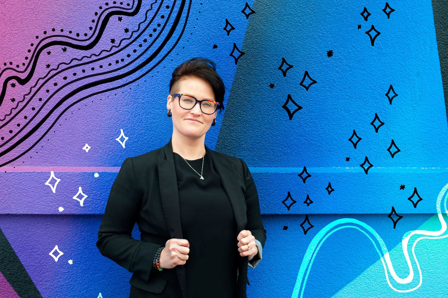 Woman in black outfit with glasses posing before colorful abstract mural wall.