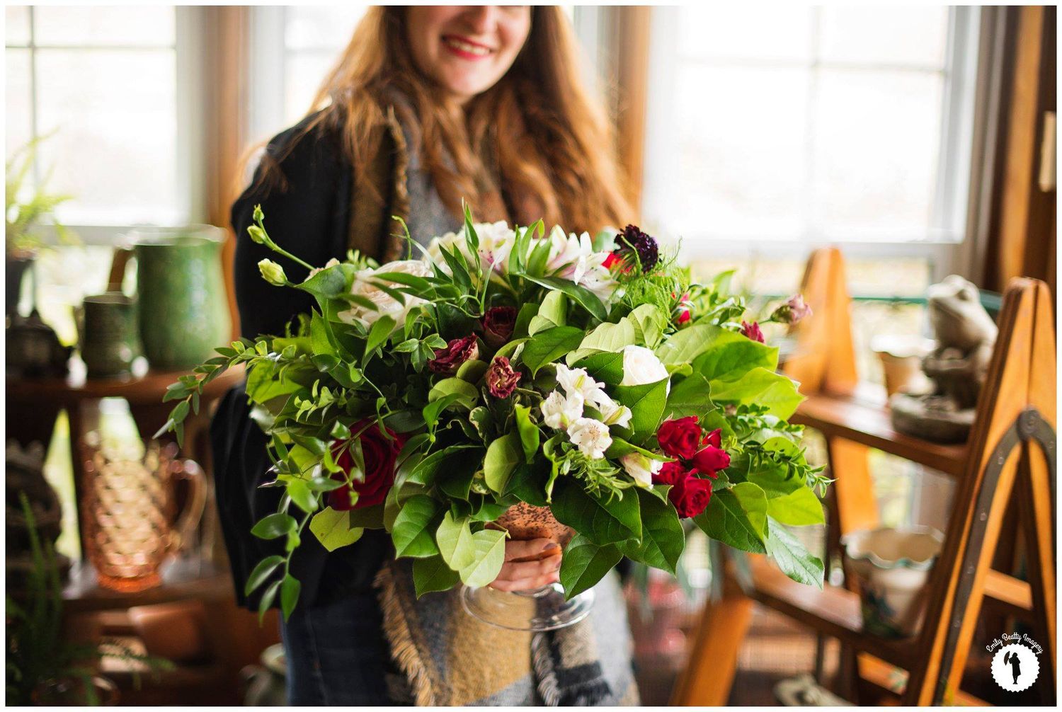 Smiling woman holding a large lush green bouquet with white and red flowers indoors near a window.