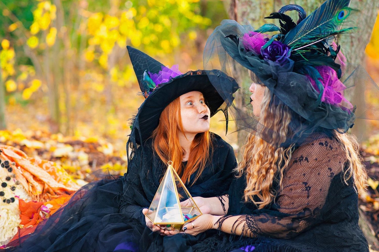 Two girls in witch costumes holding crystal ball in autumn forest during Halloween.