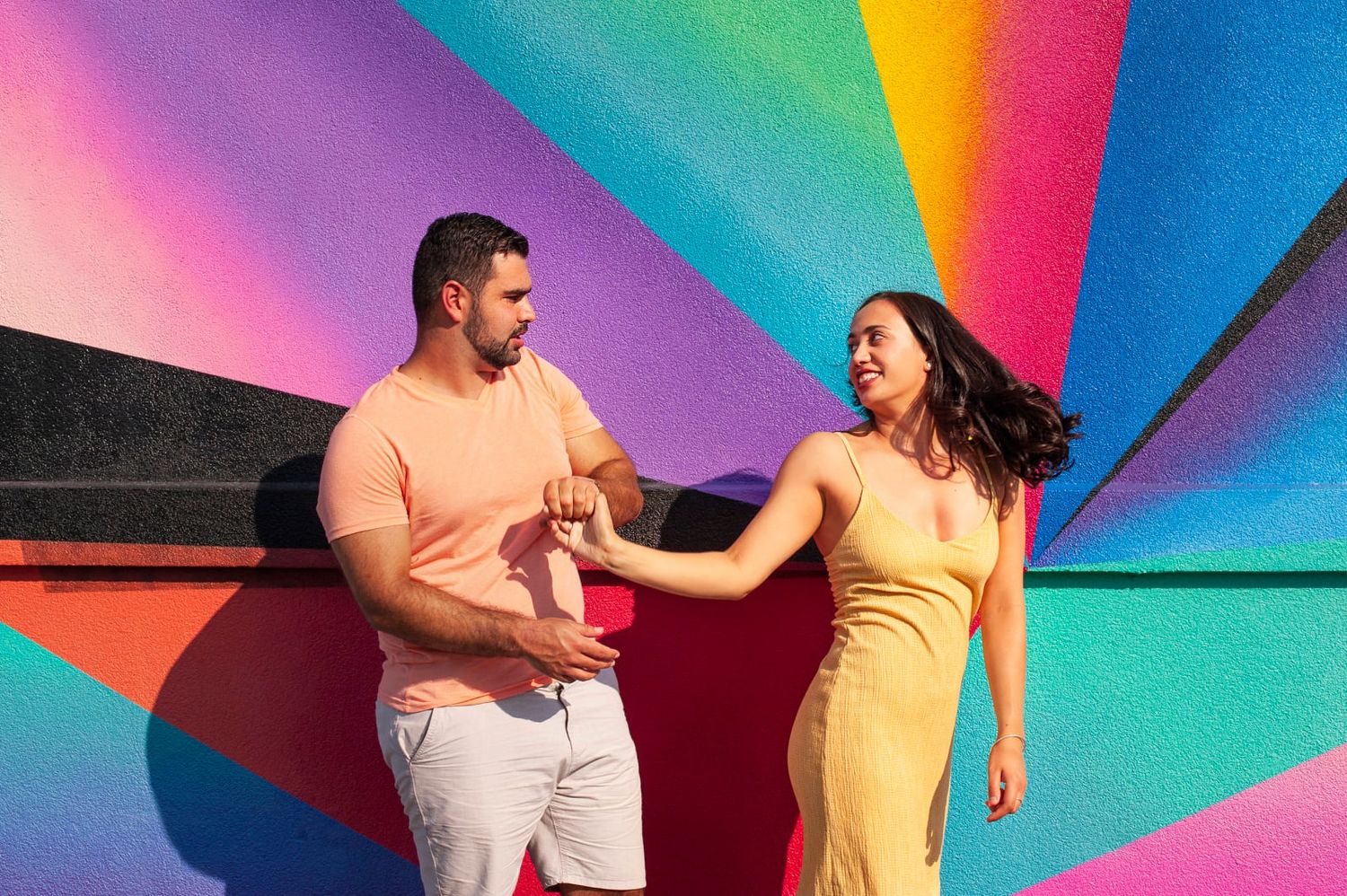 Couple dancing and holding hands in front of colorful rainbow mural wall outdoors.