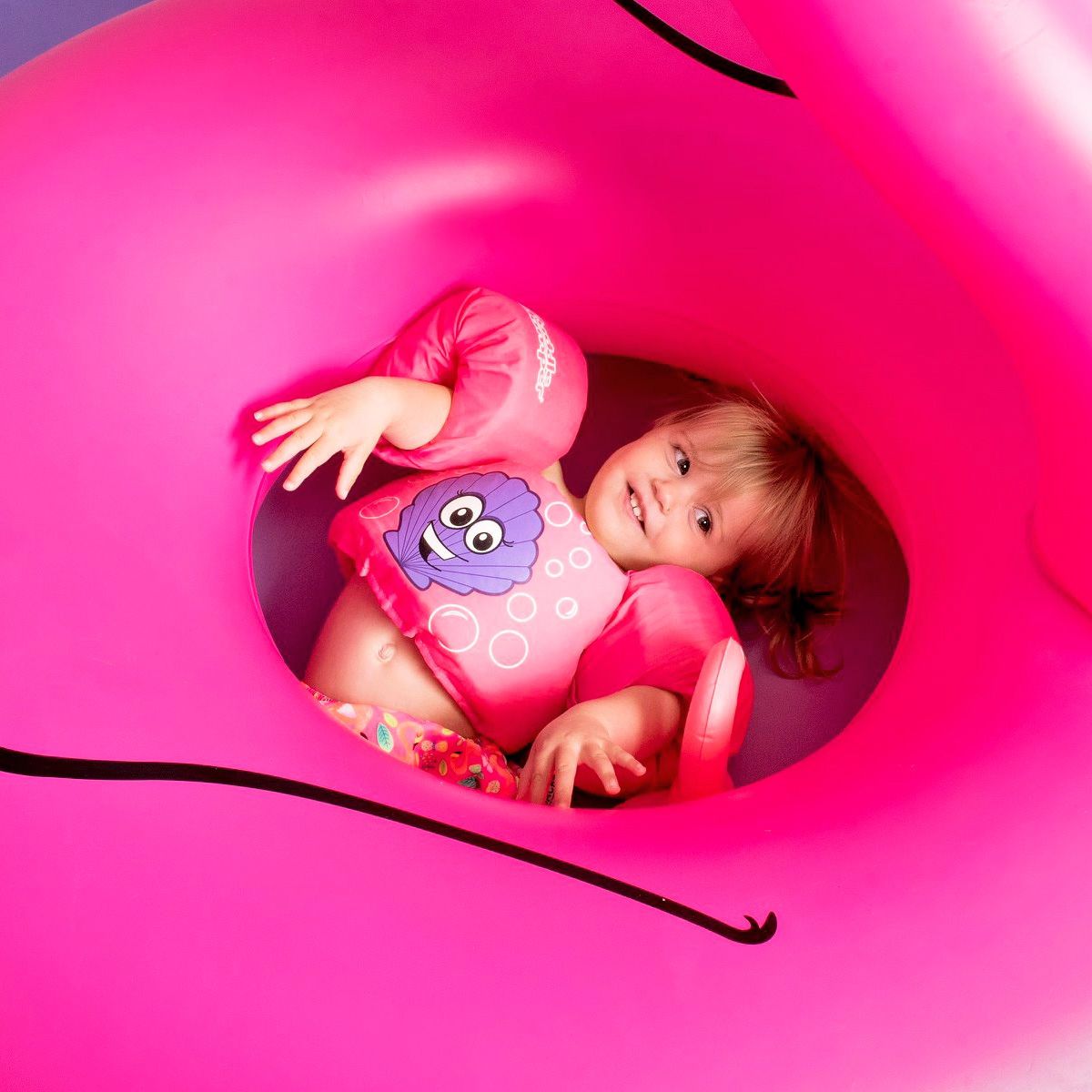 Toddler girl in pink swimsuit with floaties sits inside a large bright pink inflatable pool float ring.