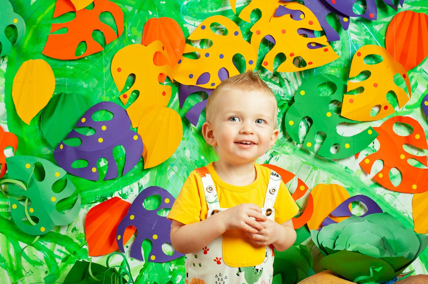Happy toddler boy in yellow shirt sitting among colorful paper tropical leaf decorations.