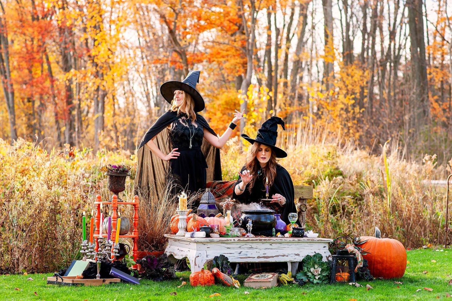 Two women in witch costumes with black hats stand at decorated outdoor table with cauldron in fall foliage.