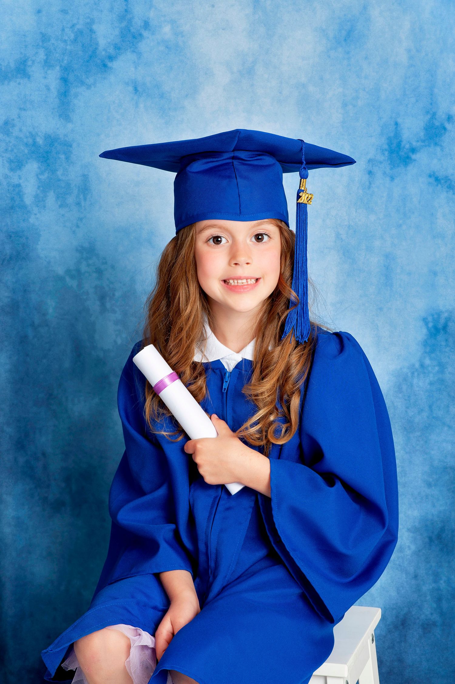 Young girl in blue graduation cap and gown holding diploma, smiling against blue mottled background.