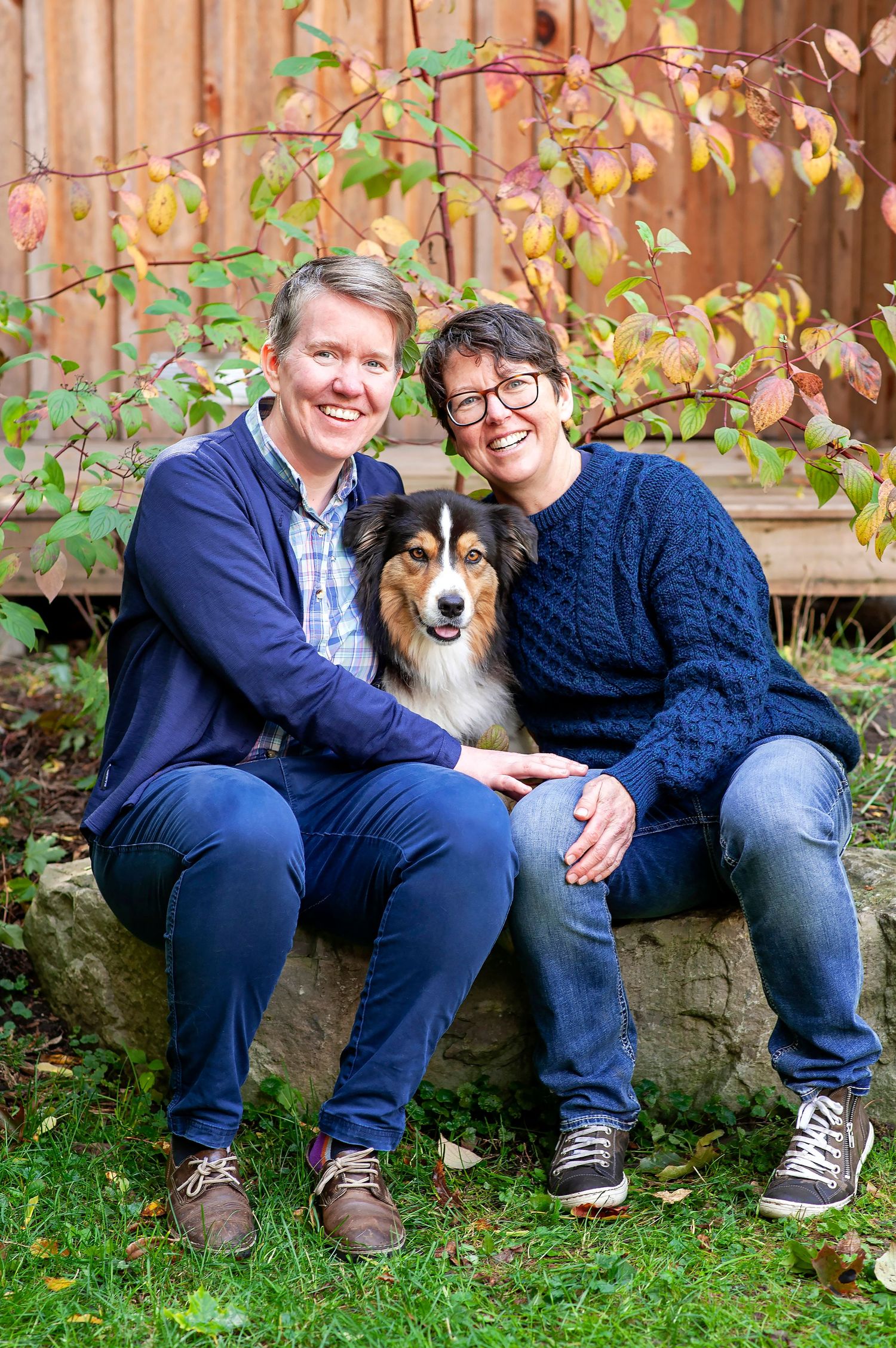 Smiling couple sitting outdoors with their Sheltie dog in autumn garden.