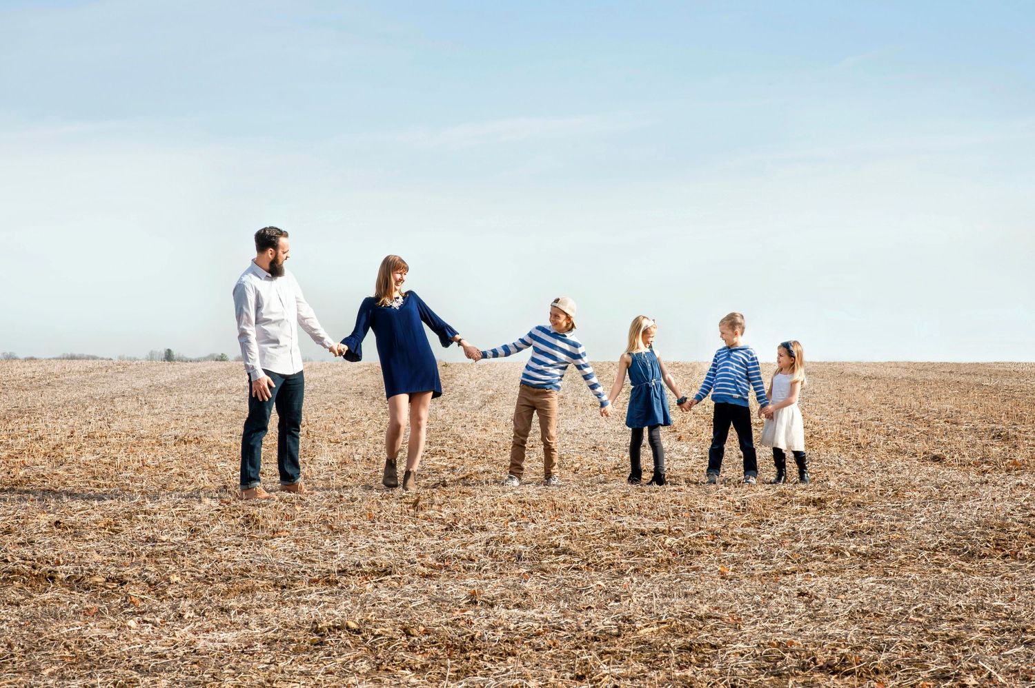 Happy family of six holding hands in a line across a golden harvested field under a blue sky.